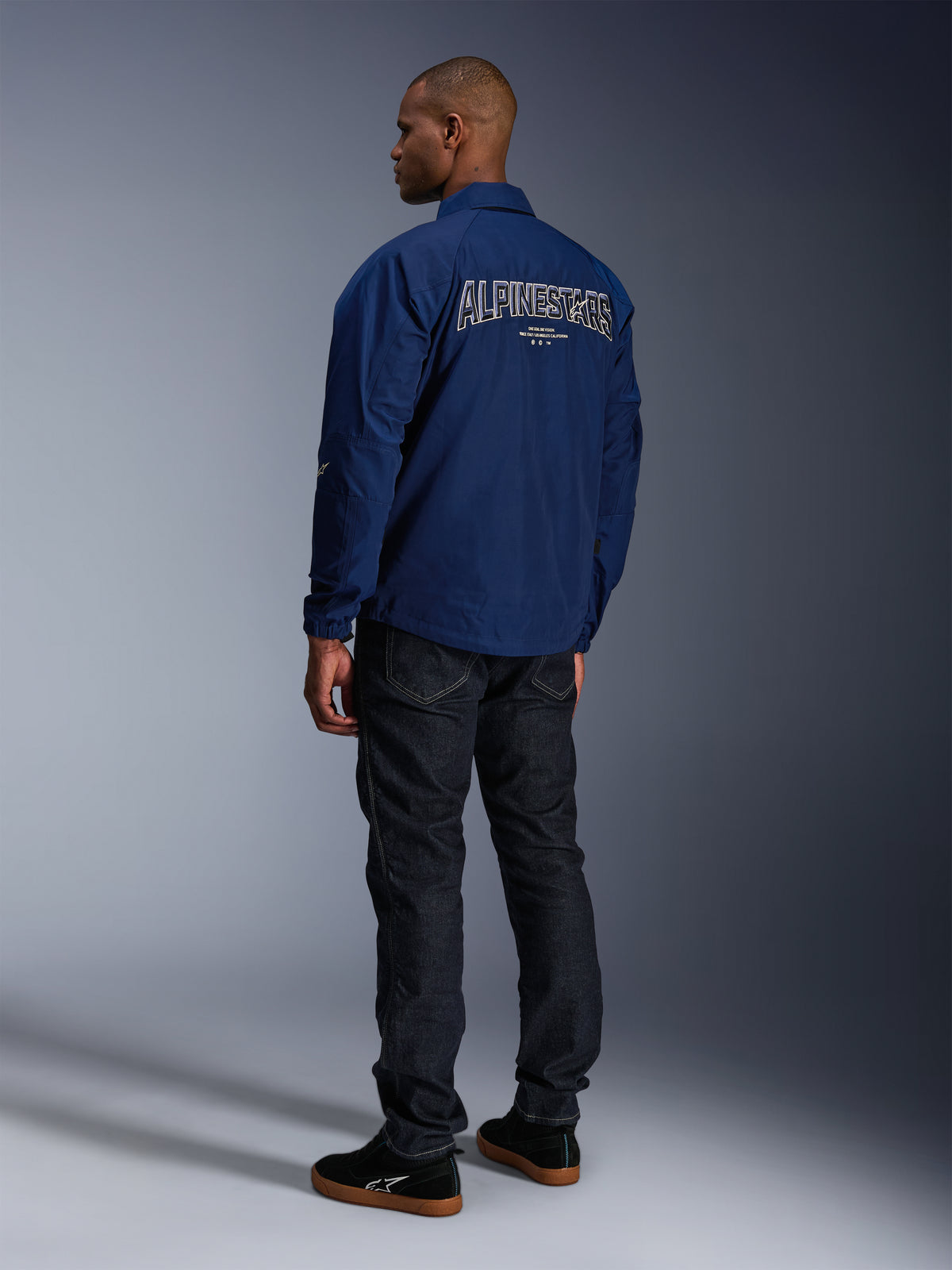 Modelo a usar o Casaco Alpinestars Revved WR em azul-marinho, visto de costas, com grande bordado 'ALPINESTARS' contornado a branco nas costas, calças de ganga escuras e ténis pretos, estética urbana de motociclismo