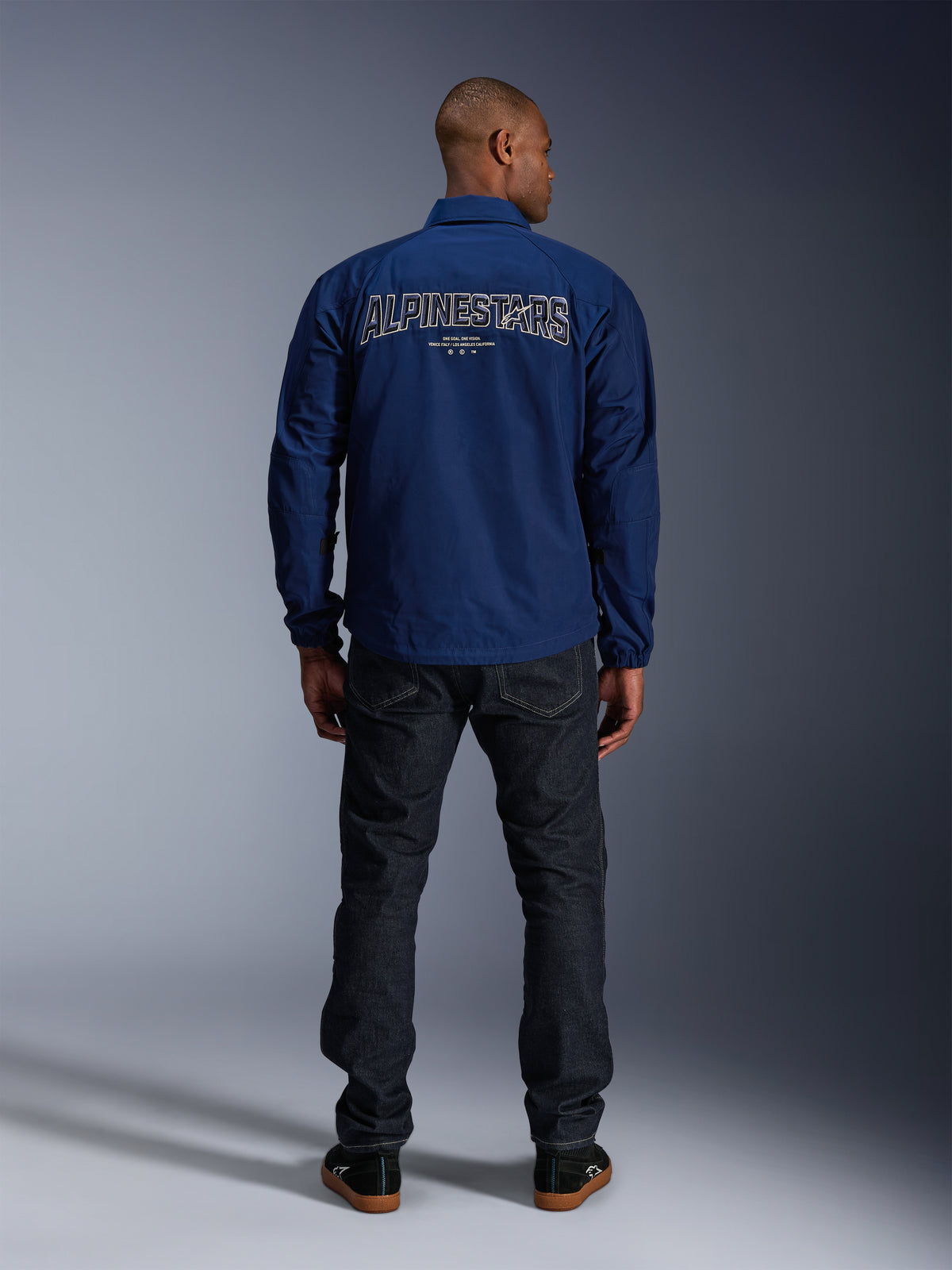 Alpinestars Revved WR Jacket em azul-marinho, vista traseira, com um grande bordado 'ALPINESTARS' com efeito 3D nas costas, combinado com calças de ganga escuras e sapatilhas pretas, vestuário urbano para motociclista