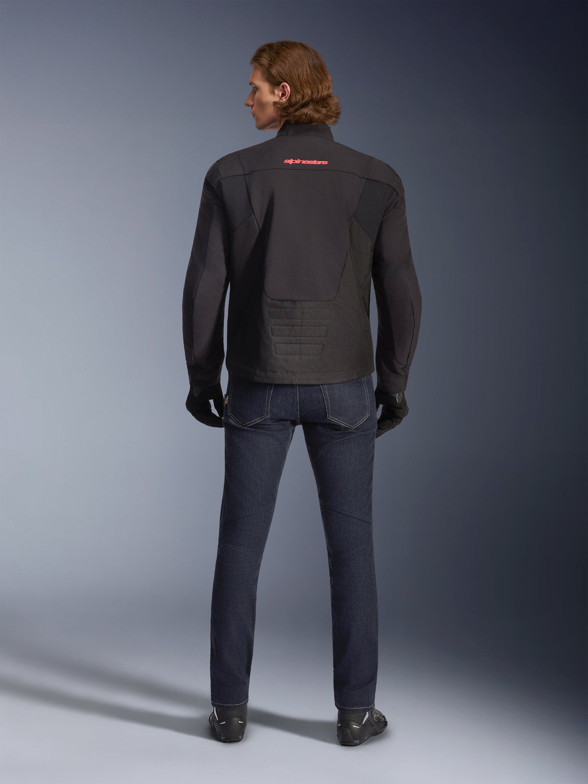 Casaco Alpinestars SMX Waterproof, preto, vista traseira num modelo, com um logótipo vermelho na parte superior das costas, bainha traseira alongada para maior cobertura, combinado com calças de ganga de motociclismo azul escuro e luvas pretas