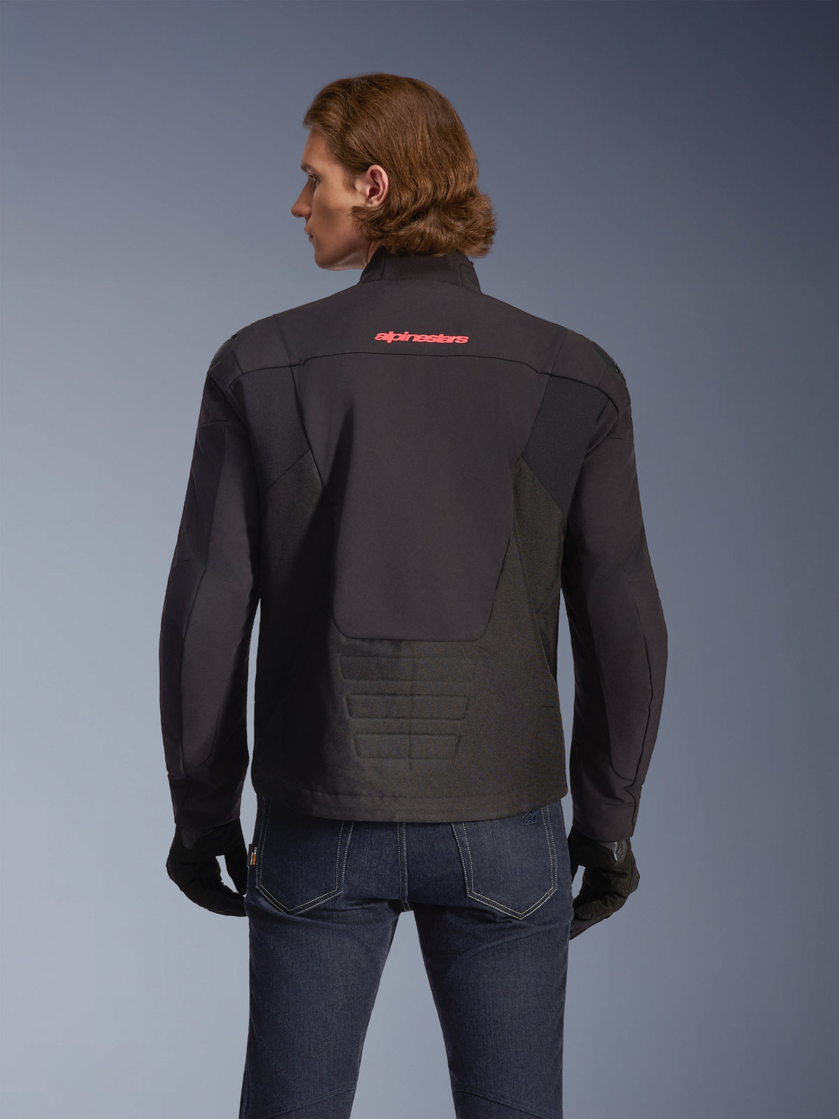 Casaco Alpinestars SMX Waterproof, preto com logótipo vermelho brilhante, vista traseira num modelo, usado com calças de ganga azul-escuro e luvas pretas, casaco têxtil de motociclismo para condução em estrada e desportiva.