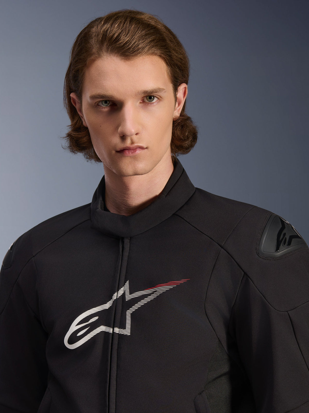 Modelo a usar o Casaco Impermeável Alpinestars SMX, preto com detalhes em branco e vermelho brilhante, com um grande logótipo de estrela branco com um traço vermelho no peito, protetores de ombro DFS Lite e uma construção têxtil elegante para desempenho em motociclismo de estrada.