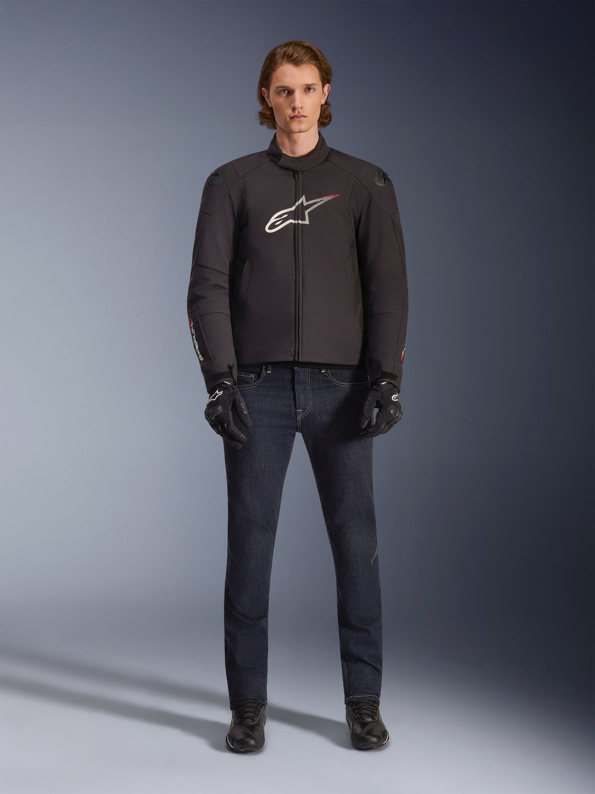 Modelo a usar o Casaco Impermeável Alpinestars SMX em preto com logótipo branco e vermelho, combinado com calças de ganga escuras para motociclismo, luvas pretas e botas de condução, conjunto de equipamento de motociclismo desportivo para todas as condições climatéricas.