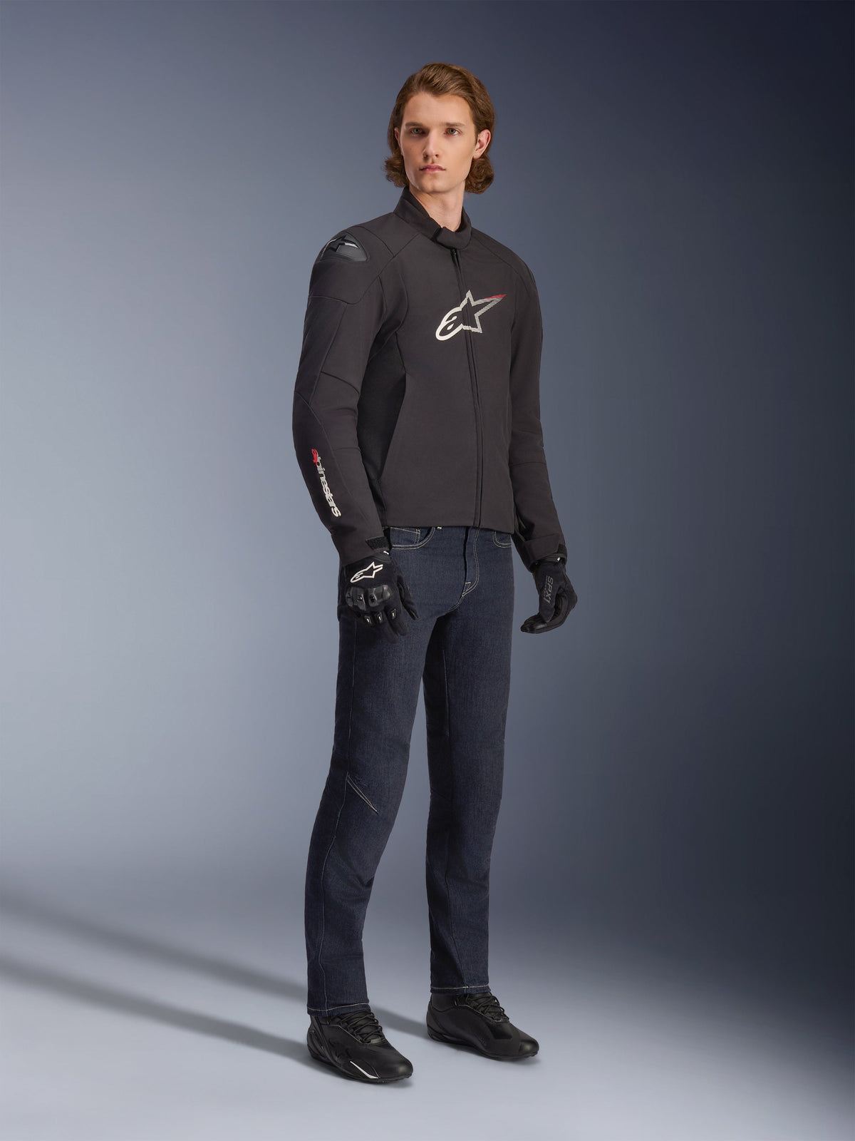 Modelo masculino a usar o Casaco Alpinestars SMX Waterproof, preto com detalhes do logótipo em branco e vermelho, combinado com calças de ganga azul escuro e luvas de motociclismo pretas, casaco de motociclismo têxtil com corte desportivo elegante
