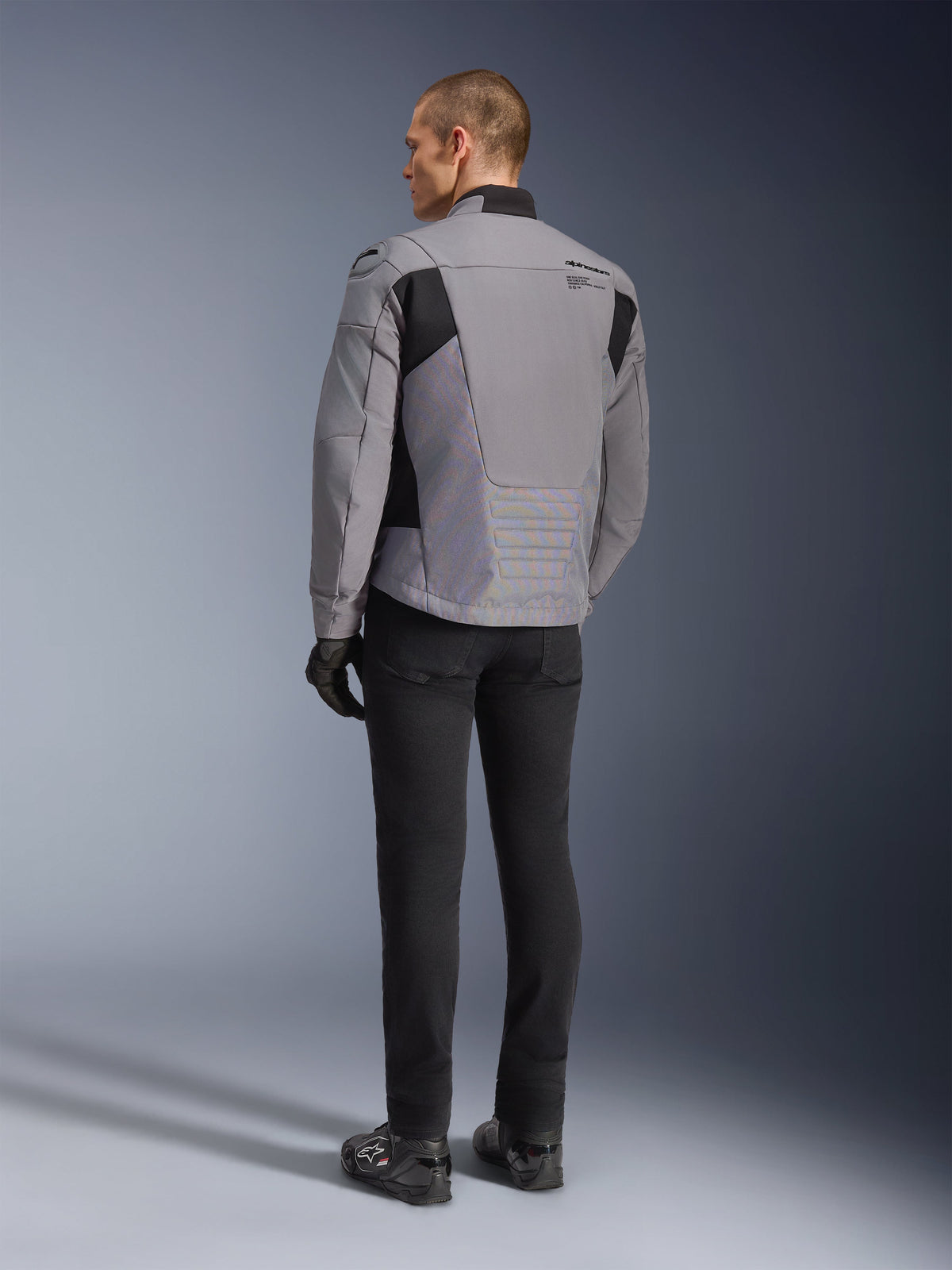 Casaco Impermeável Alpinestars SMX em cinzento escuro e preto, vista traseira num modelo, usado com calças e botas pretas, com construção em tecido técnico, painéis pretos e logótipo Alpinestars no ombro.