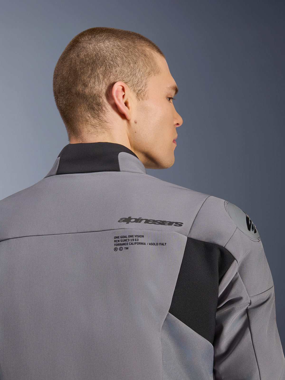 Casaco Alpinestars SMX Waterproof em cinzento escuro e preto, grande plano da zona das costas e dos ombros mostrando o logótipo Alpinestars e a proteção de ombro DFS lite, vestido por um modelo masculino