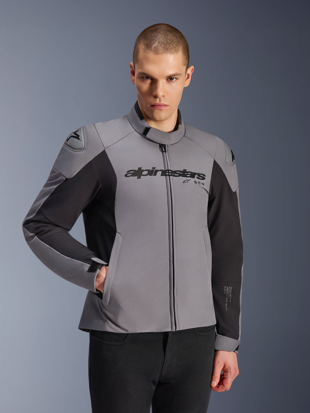 Casaco Alpinestars SMX Waterproof, cinzento escuro e preto, vestido por um modelo masculino, com a marca 'alpinestars' no peito, proteção de ombros DFS Lite, design desportivo elegante com materiais têxteis e softshell para condução em estrada.