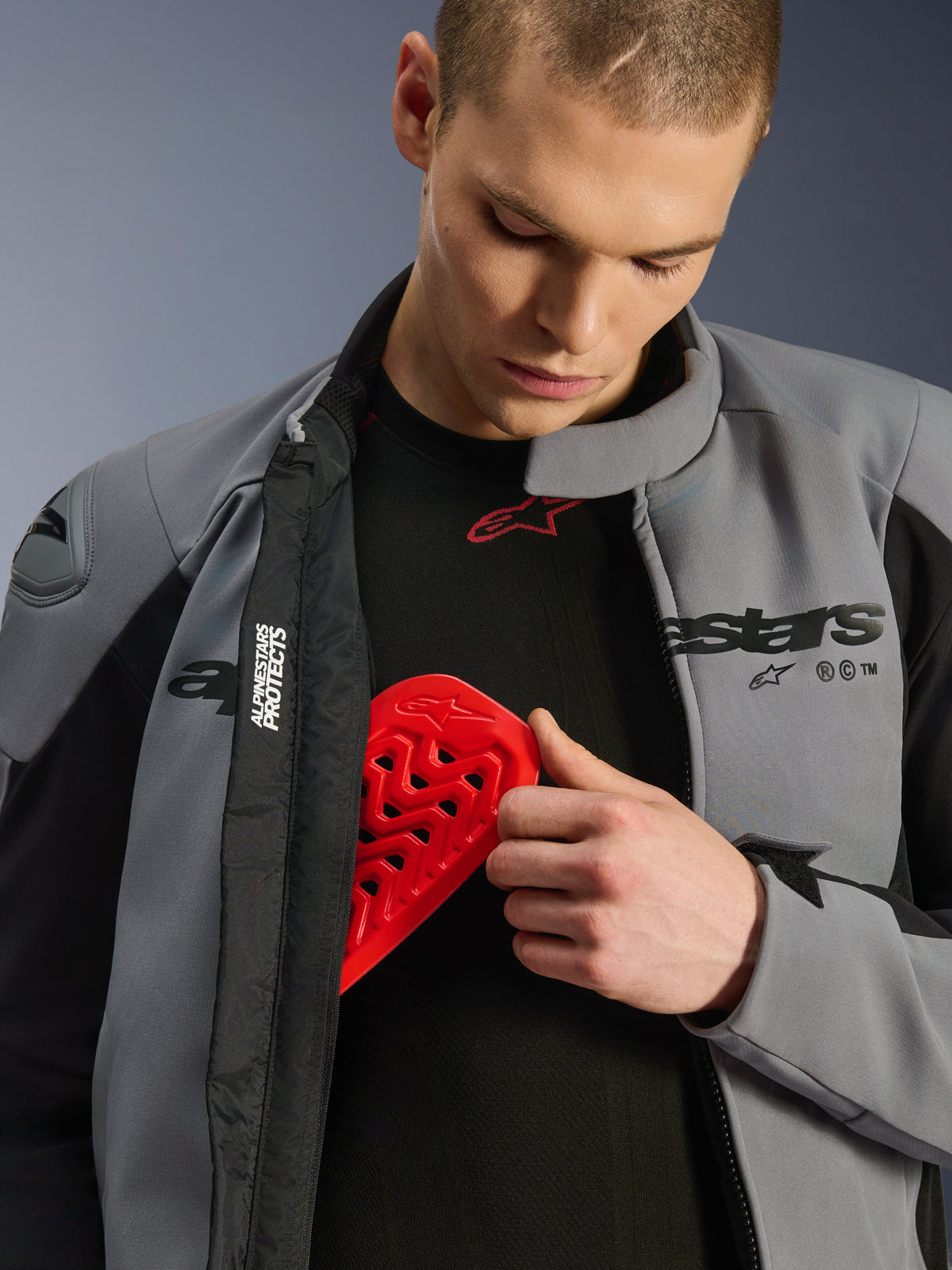 Modelo masculino a usar o Casaco Alpinestars SMX Waterproof em cinzento escuro e preto, a inserir um protetor de peito Nucleon vermelho num bolso interno, demonstrando a compatibilidade do casaco com proteções e as suas características de proteção.