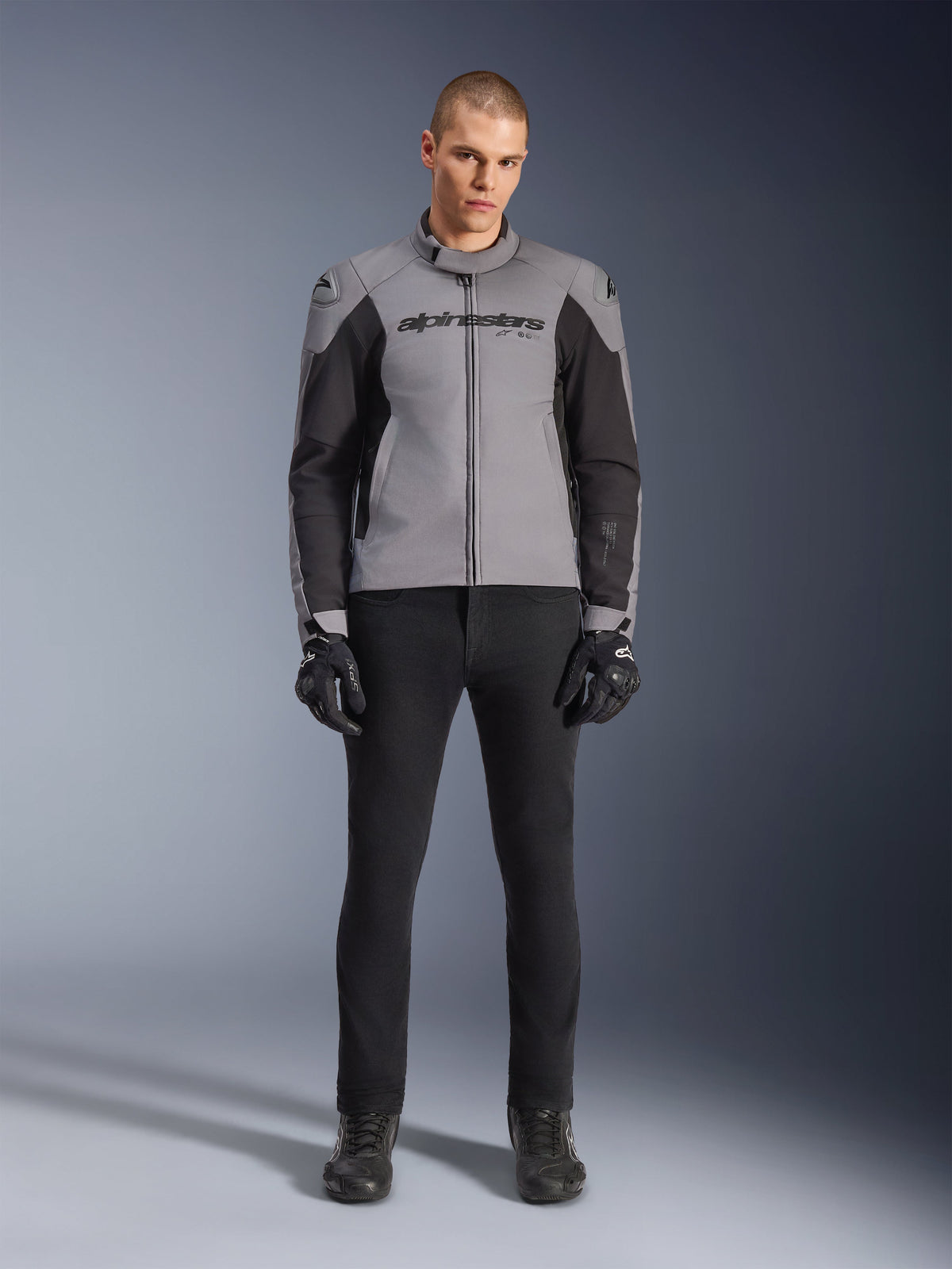 Casaco Alpinestars SMX Waterproof, cinzento escuro e preto, vista frontal num modelo masculino, casaco têxtil desportivo de motociclismo com painéis laterais pretos e marca Alpinestars no peito, combinado com calças e luvas de motociclismo pretas.