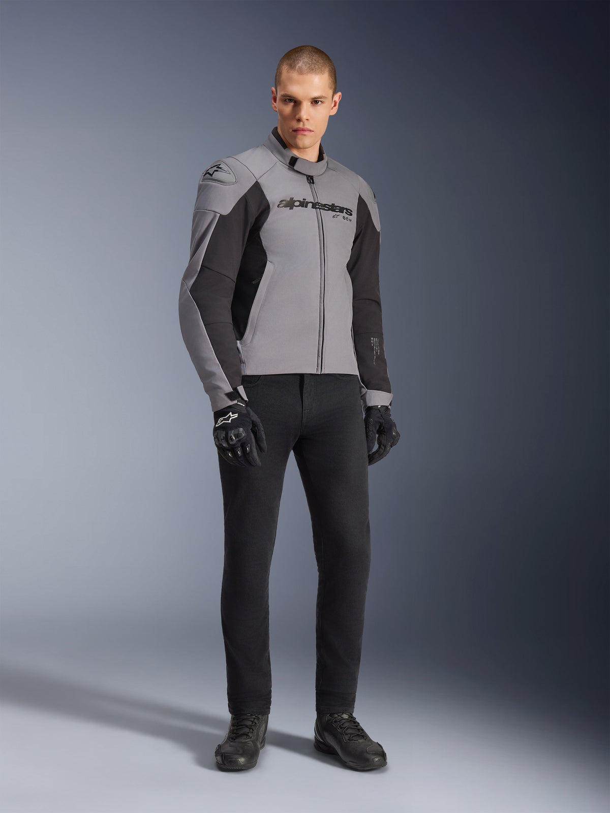Alpinestars SMX Waterproof Jacket, casaco de motociclismo, cinzento escuro e preto, usado por modelo com luvas de condução pretas e calças pretas, logótipo proeminente no peito e design técnico desportivo para condução em estrada em todas as condições climatéricas