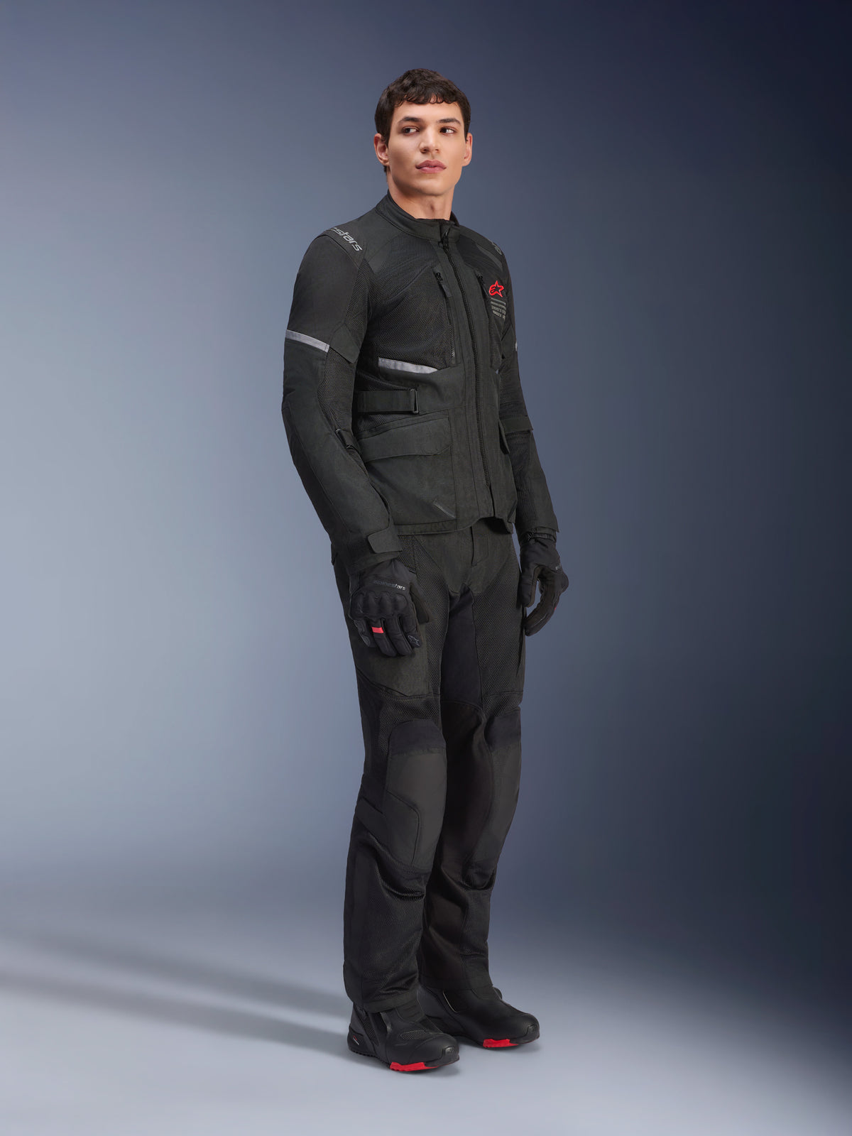 Casaco Alpinestars Andes Air Drystar®, preto, vestido por um modelo como parte de um conjunto completo de aventura para motociclismo com calças, luvas e botas a condizer, apresentando painéis de ventilação em malha, logótipo vermelho no peito e faixas refletoras para visibilidade em touring.