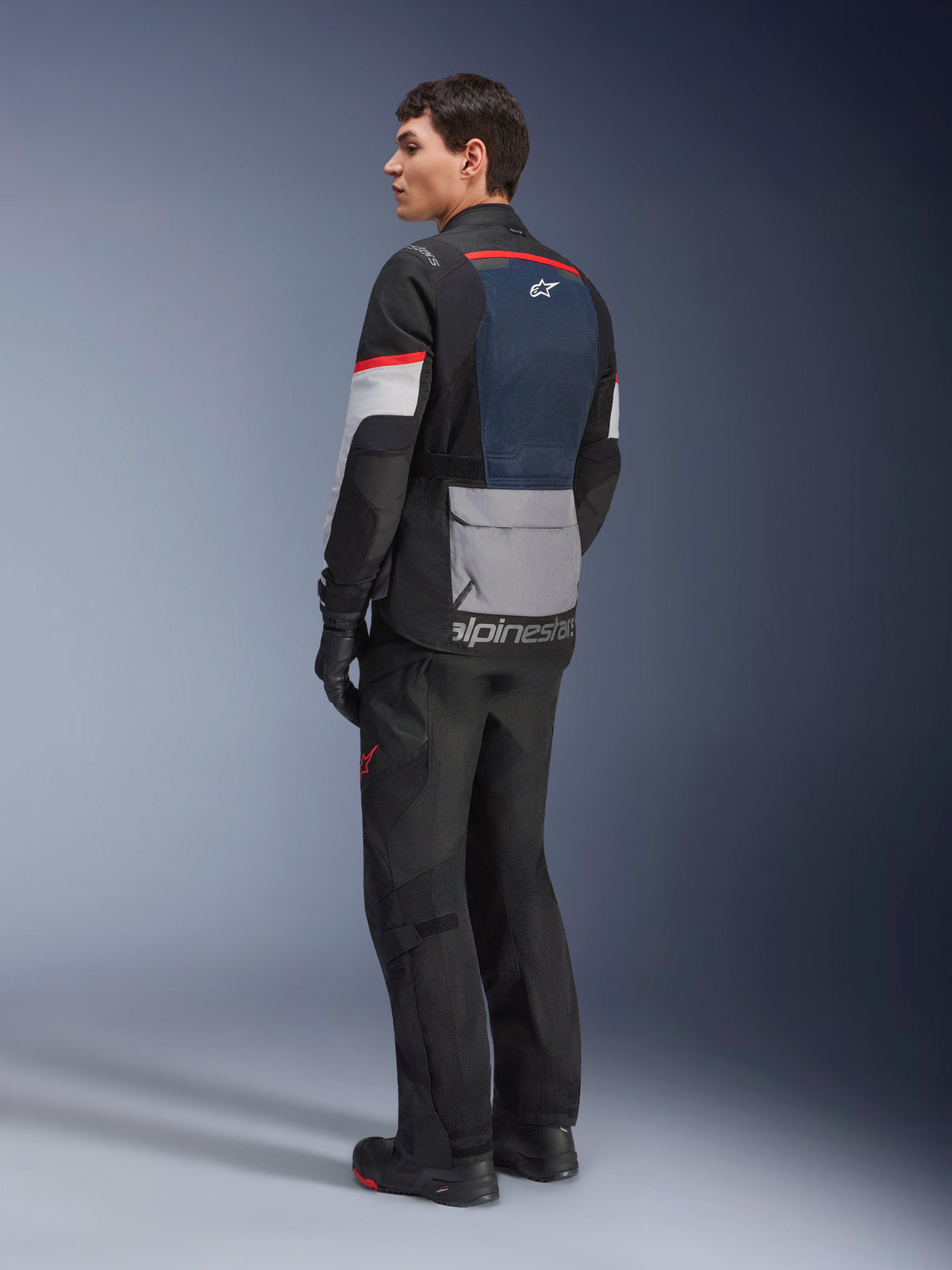Male model from a rear-side view wearing the Alpinestars Andes Air Drystar® Jacket in Deep Blue Black Ice Gray, paired with black motorcycle pants and gloves, featuring a blue mesh back panel, gray utility pocket on the lower back, and red accents on the sleeves, professional adventure touring gear.