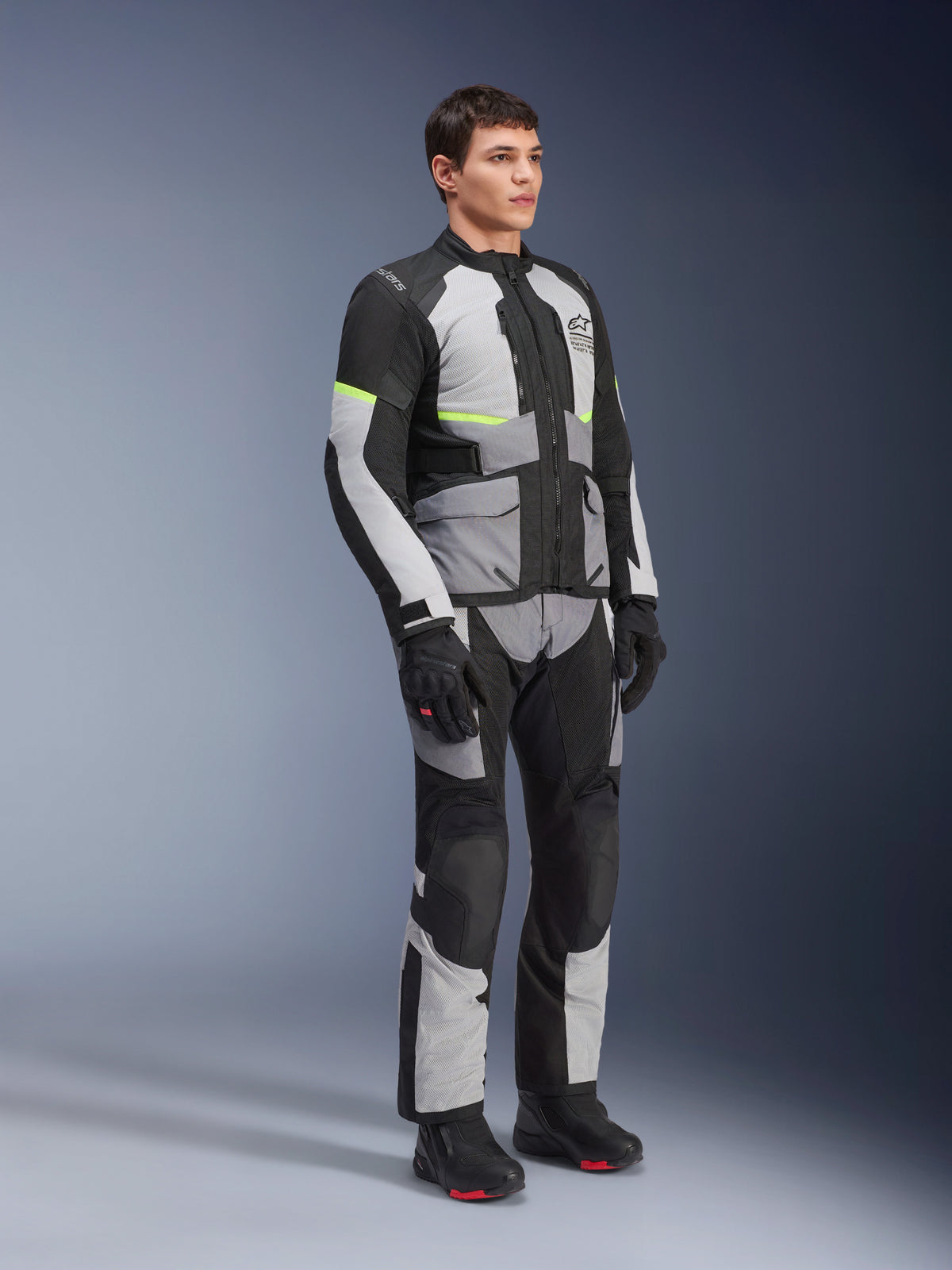 Casaco Alpinestars Andes Air Drystar®, em cinzento gelo, cinzento escuro e preto, vestido por um modelo num conjunto completo de touring de aventura com calças a condizer e botas pretas, com ventilação em malha, detalhes néon e logótipo Alpinestars.