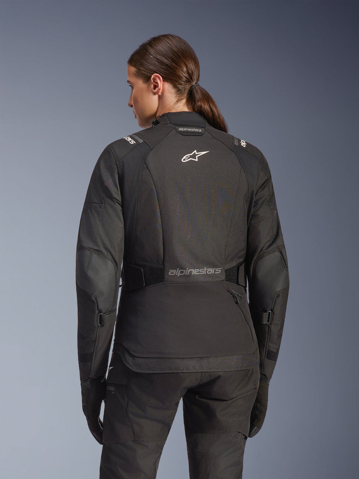Woman wearing Alpinestars Stella Andes V4 Drystar Jacket in Black, rear view featuring white logo on the back and reinforced elbow panels, paired with matching black pants and gloves, adventure touring motorcycle outfit