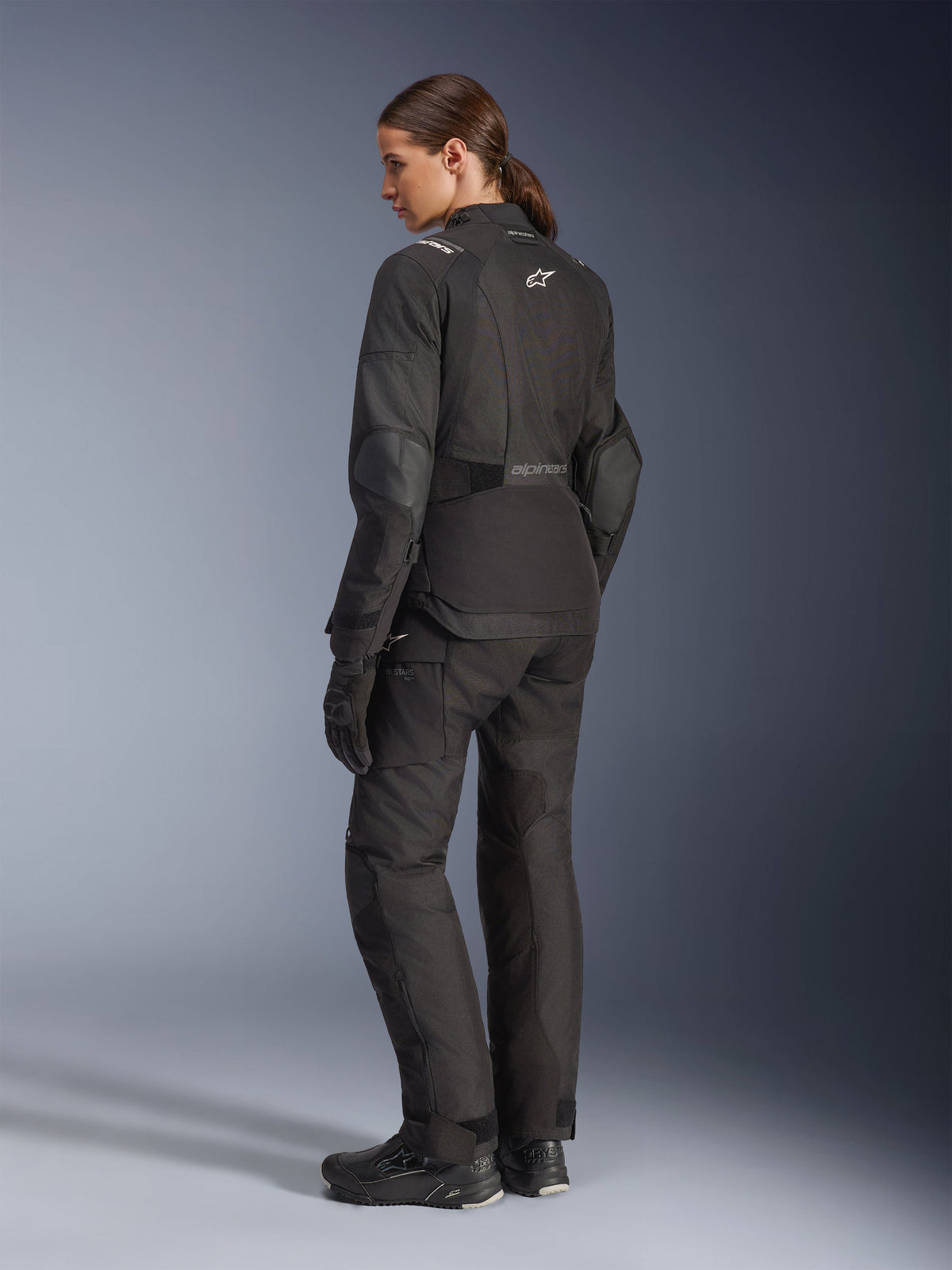Woman rider wearing Alpinestars Stella Andes V4 Drystar® Jacket, Black adventure motorcycle jacket, 3/4 rear view, paired with matching textile pants and gloves, featuring white Alpinestars logos on the back and waist, reinforced protective panels, and an elongated rear hem.