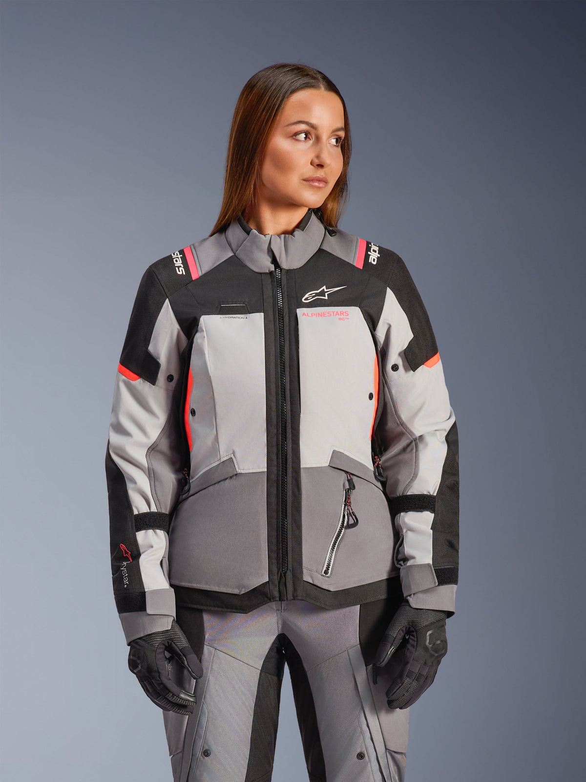 Alpinestars Stella Andes V4 Drystar, casaco de moto para mulher, cinzento e preto com detalhes coral, impermeável, modelo a usar equipamento têxtil de aventura.
