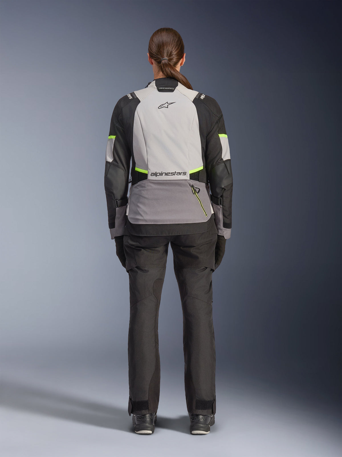 Woman rider from the back wearing the Alpinestars Stella Andes V4 Drystar® Jacket, Ice Gray, Dark Gray, and Black with Yellow Fluo accents, featuring a large rear storage pocket and Alpinestars logo, paired with matching black adventure pants, all-weather motorcycle touring gear