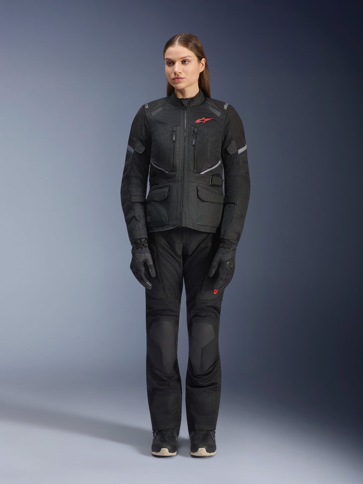 Modelo feminina a usar o casaco Alpinestars Stella Andes Air Drystar® em preto, combinado com calças de turismo e luvas pretas a condizer, com grandes painéis de ventilação em malha, detalhes do logótipo em vermelho e elementos refletores, fotografia profissional de estúdio de equipamento de motociclismo para mulher.