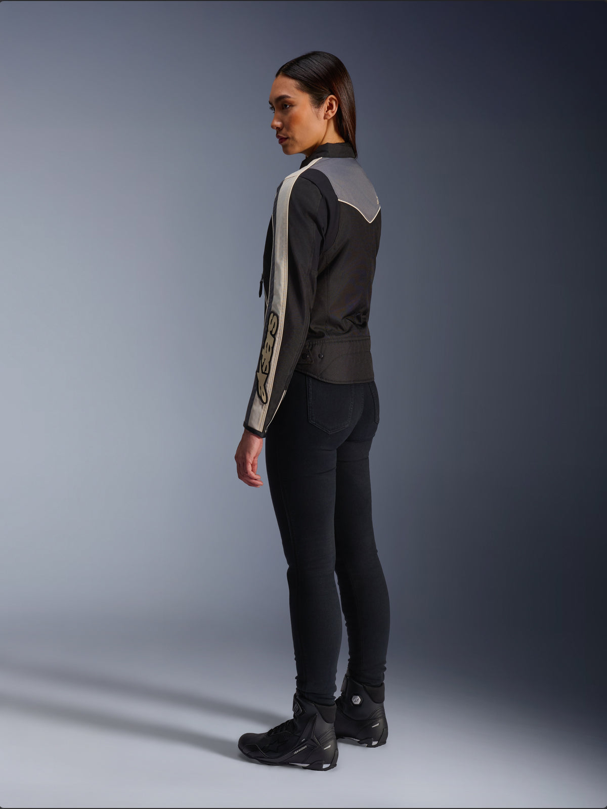 Modelo feminina vista de lado e de costas a usar o Casaco Alpinestars Stella T-Dyno WR em Preto Cinzento Escuro Ecru, com um design de inspiração vintage, riscas ecru nas mangas e um painel traseiro cinzento, combinado com calças pretas e calçado de motociclismo preto.