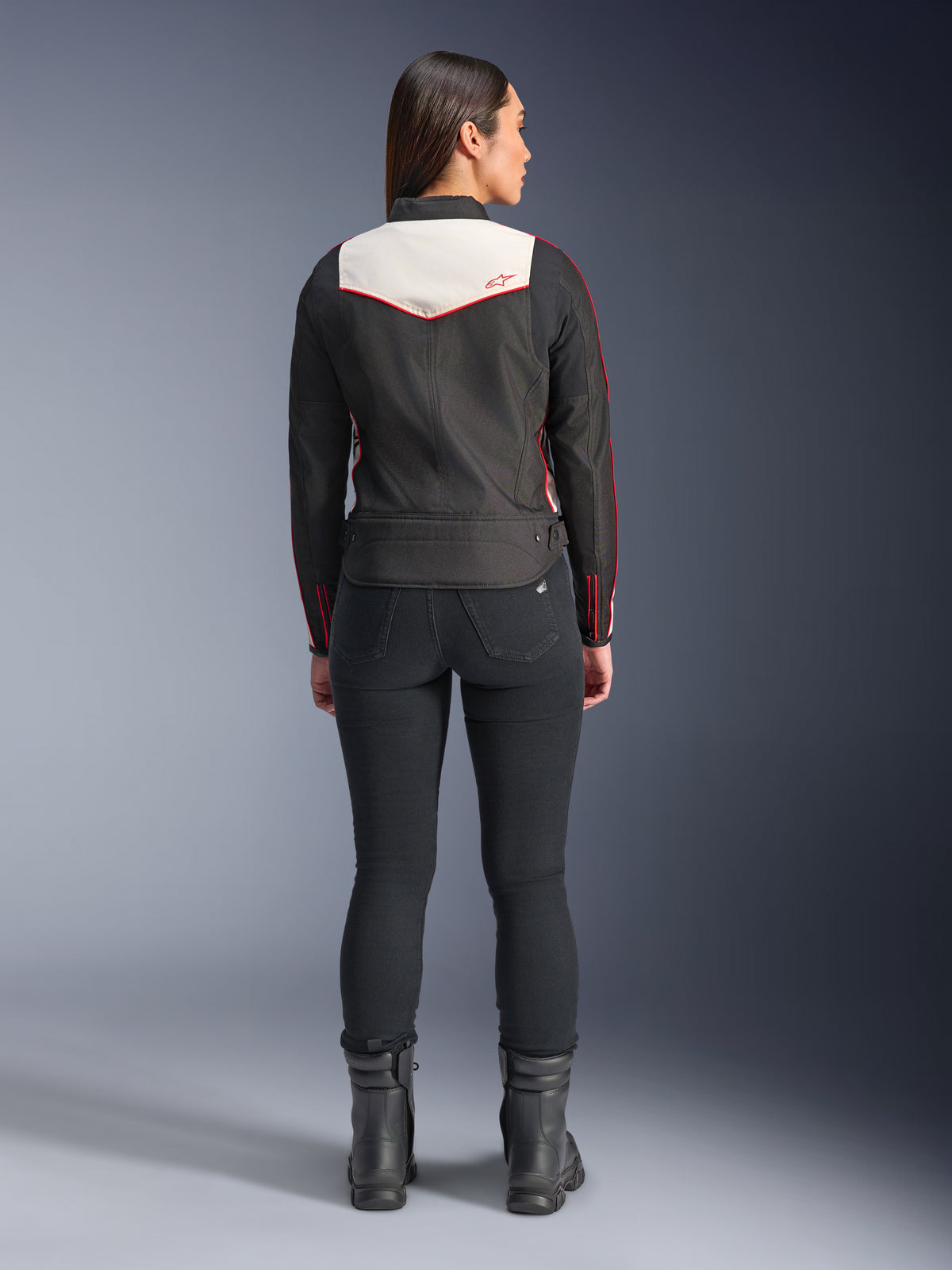 Vista traseira de uma modelo feminina a usar o casaco Alpinestars Stella T-Dyno WR em Preto, Cru e Vermelho Brilhante, com um painel traseiro creme proeminente e detalhes do logótipo em vermelho, combinado com calças e botas pretas, um casaco de motociclismo têxtil de inspiração vintage.