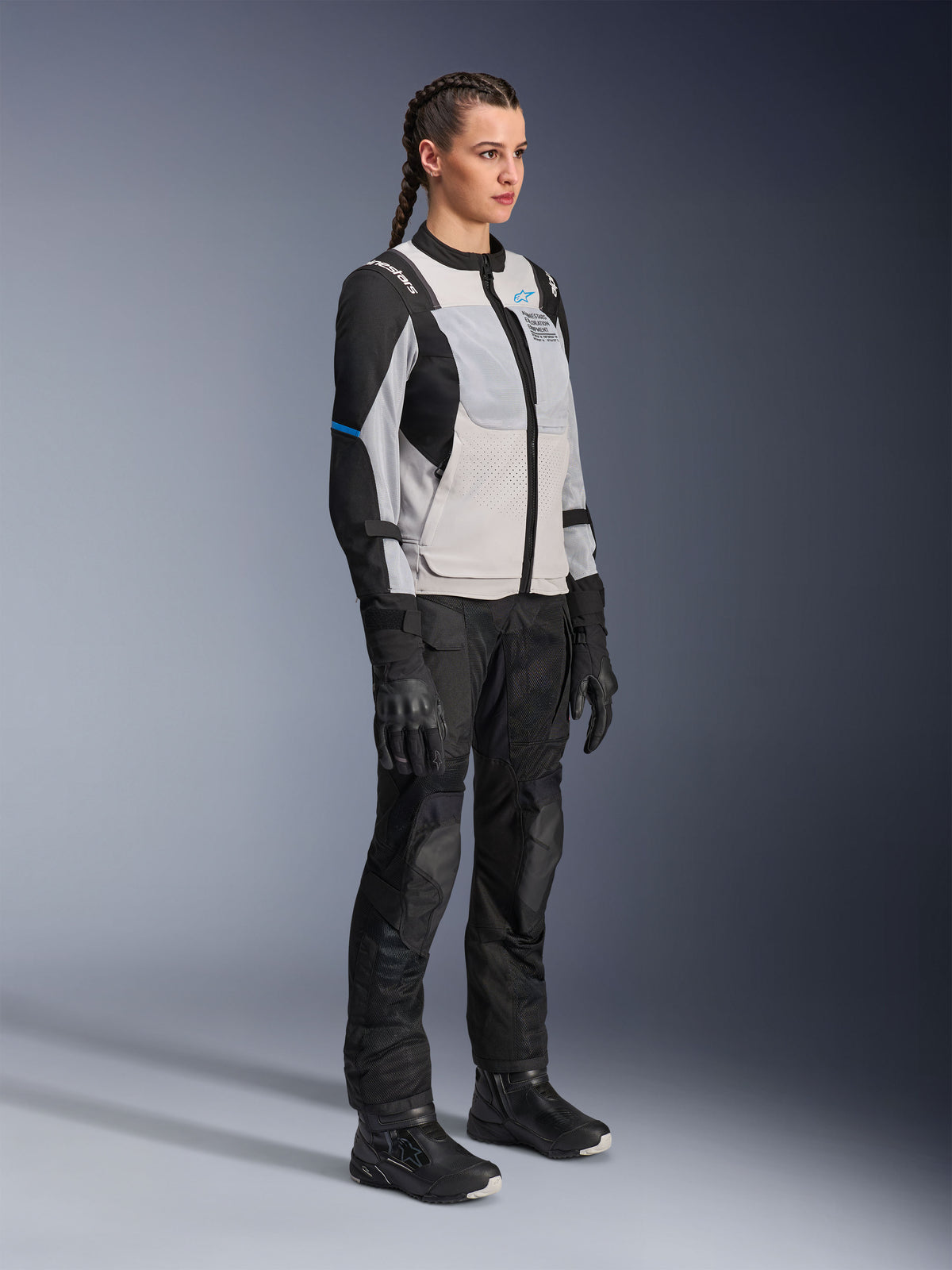 Modelo a usar o Casaco Alpinestars Stella ST-2 Air em Ice Gray e preto, casaco técnico de motociclismo para mulher com painéis de malha e construção em softshell, combinado com calças e botas de touring de aventura pretas, com detalhes em azul e um design de condução moderno