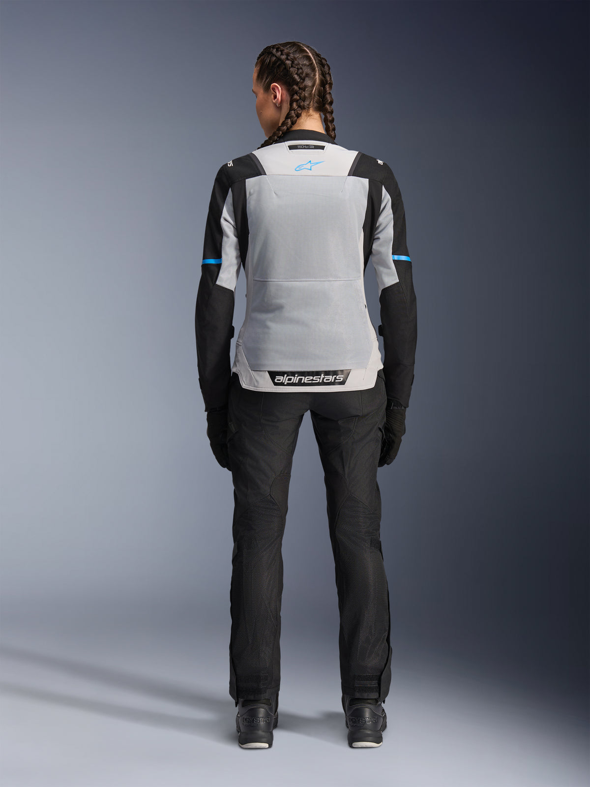 Casaco Alpinestars Stella ST-2 Air, Ice Gray Black, vista traseira em modelo feminina, com um grande painel traseiro em malha cinzento claro, mangas pretas com detalhes em azul, marca Alpinestars na zona lombar, combinado com calças técnicas e botas pretas, equipamento de touring de verão para motociclismo