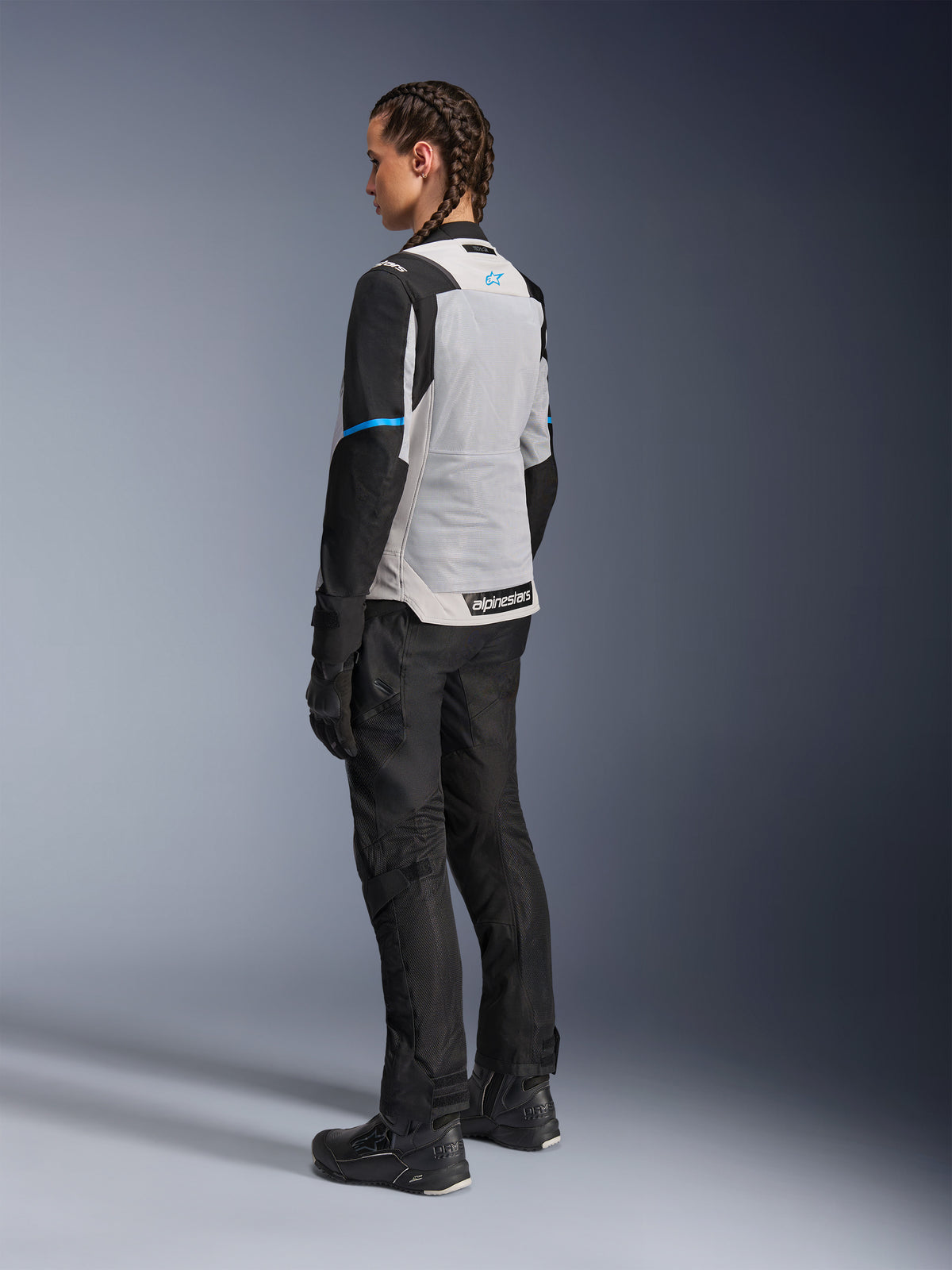 Motociclista feminina a usar o Casaco Alpinestars Stella ST-2 Air em cinzento gelo e preto, vista traseira de três quartos realçando o amplo painel de malha cinzento claro nas costas, mangas pretas com um detalhe azul e logótipo Alpinestars azul, combinado com calças, luvas e botas pretas para um equipamento completo de adventure touring.