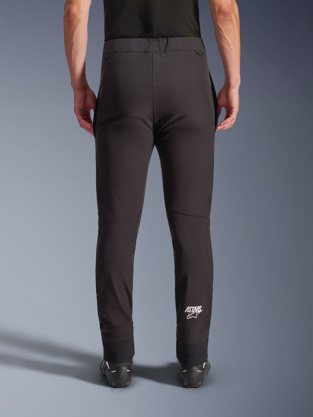 Alpinestars Aeroshell Pants, calças de motociclismo softshell pretas, vista traseira mostrando o corte justo e joelhos pré-curvados, logótipo Astars branco na parte inferior da perna direita, bainha elástica, design crossover para condução em estrada