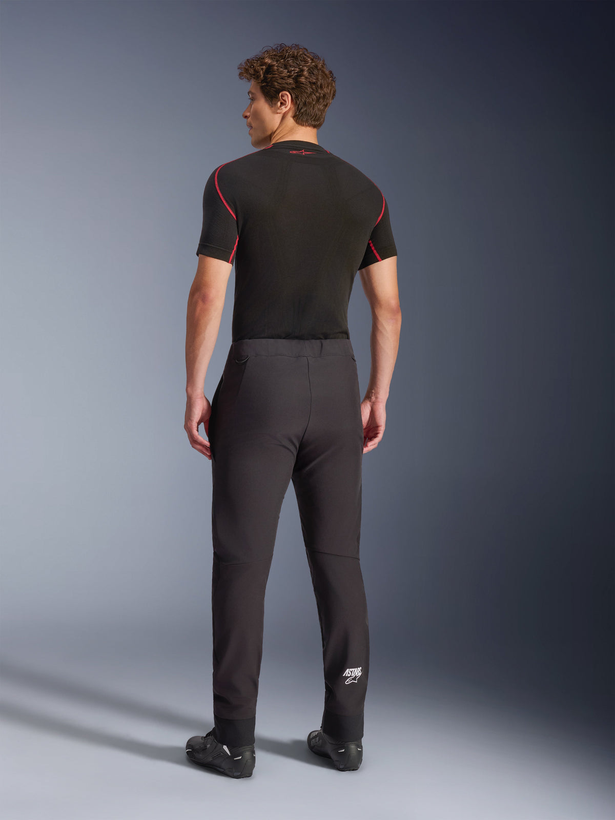 Calças Alpinestars Aeroshell, pretas, calças de motociclismo softshell, vista traseira num modelo, com um corte afunilado moderno, logótipo Astars branco na parte inferior da perna e bainhas elásticas, usadas com uma camada de base preta de manga curta e sapatos de condução.
