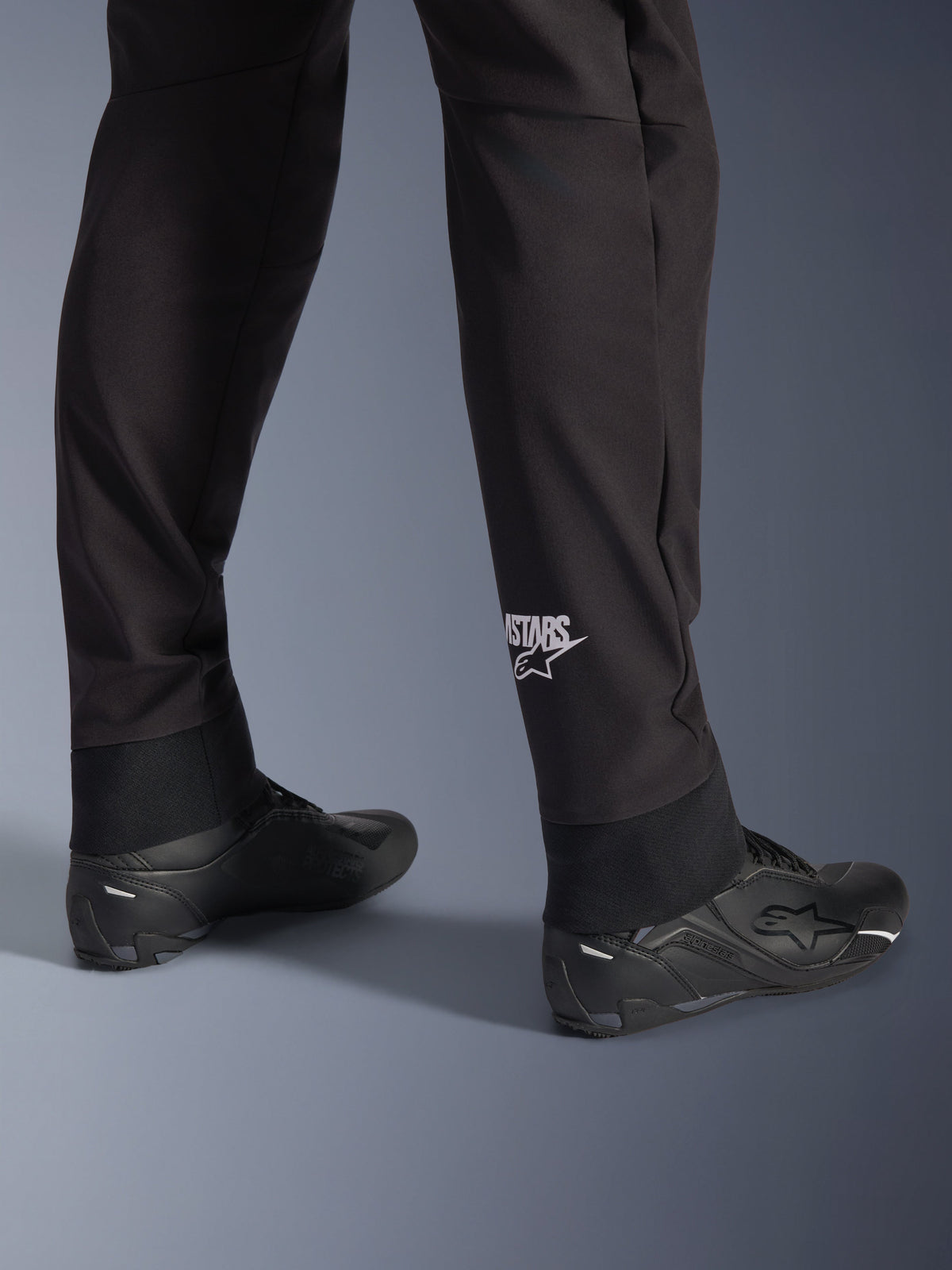 Alpinestars Aeroshell Pants, calças de motociclismo em softshell pretas, grande plano da parte inferior da perna e bainha elástica, logótipo Alpinestars branco na zona da barriga da perna, combinadas com sapatos de condução pretos, design desportivo crossover moderno