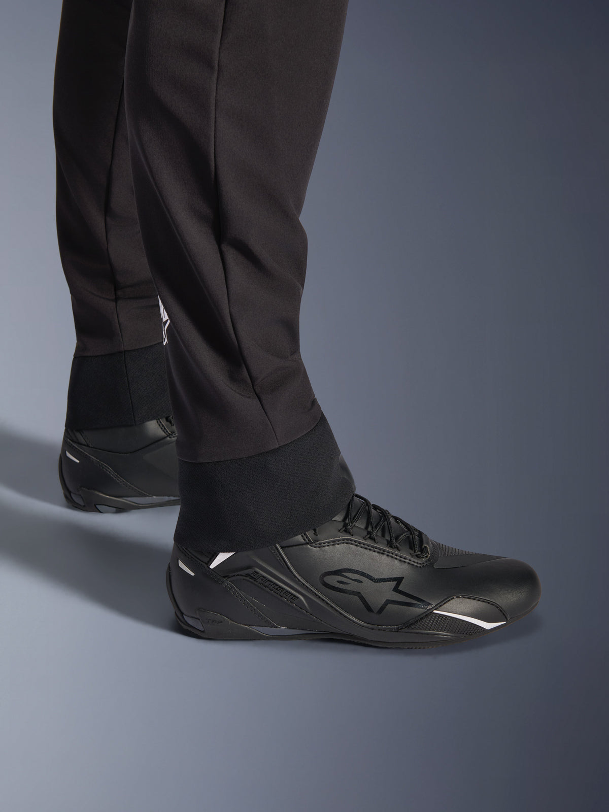 Calças Alpinestars Aeroshell, pretas, vista em pormenor da bainha inferior e punho elásticos, design moderno de softshell desportivo, usadas com sapatos de condução pretos da Alpinestars