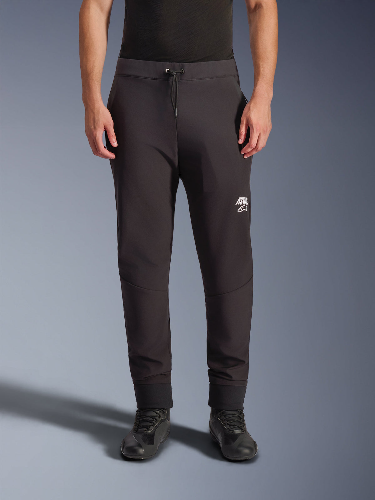 Alpinestars Aeroshell Pants, calças de motociclismo softshell pretas, corte atlético estilo jogger com cordão na cintura e punhos elásticos, com logótipo Astars branco, usadas com calçado de condução preto, equipamento crossover moderno para estrada e uso casual