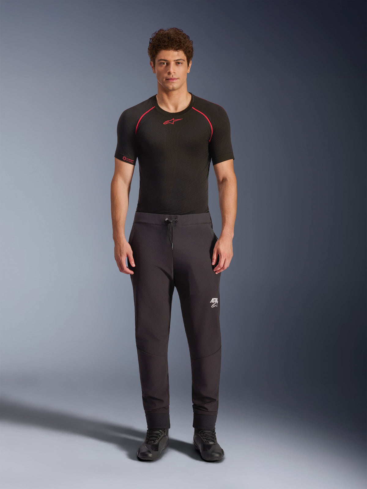 Alpinestars Aeroshell Pants, calças de motociclismo softshell pretas, usadas com uma camisola interior de manga curta preta e sapatos pretos, com cintura elástica com cordão, logótipo Astars branco na coxa e bainhas elásticas, silhueta atlética moderna para condução em estrada
