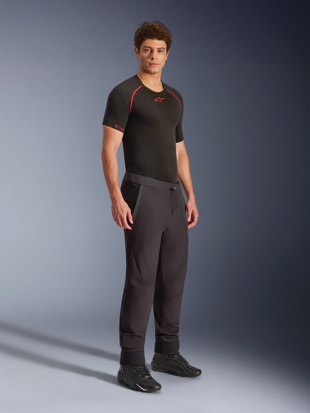 Alpinestars Aeroshell Pants, pretas, calças de motociclismo softshell desportivas crossover, usadas por um modelo masculino com uma camada de base técnica preta e sapatos pretos, com cintura com cordão e bainhas elásticas.