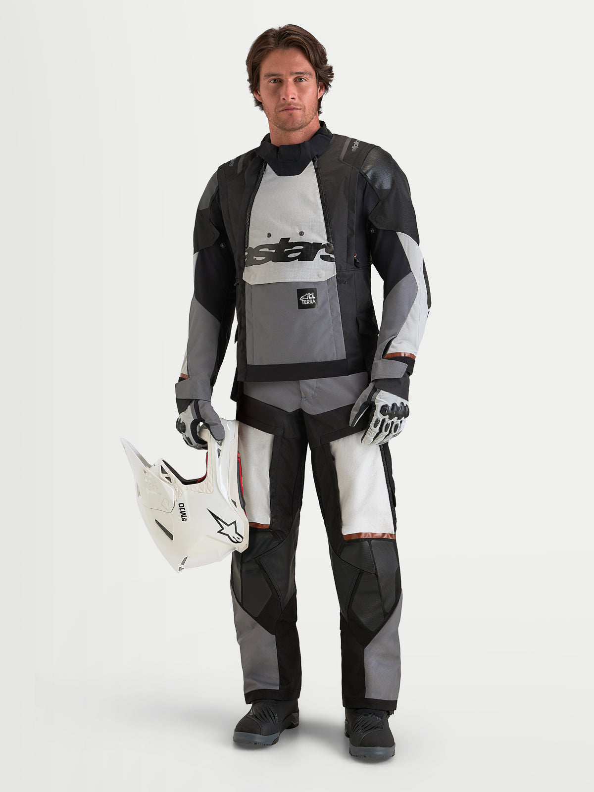 Alpinestars Harok V2 Drystar, calças de moto de aventura para homem em cinzento-escuro, cinzento-gelo e preto, vestidas num modelo com casaco ADV a condizer e a segurar um capacete branco, equipamento de proteção certificado CE para todo-o-terreno