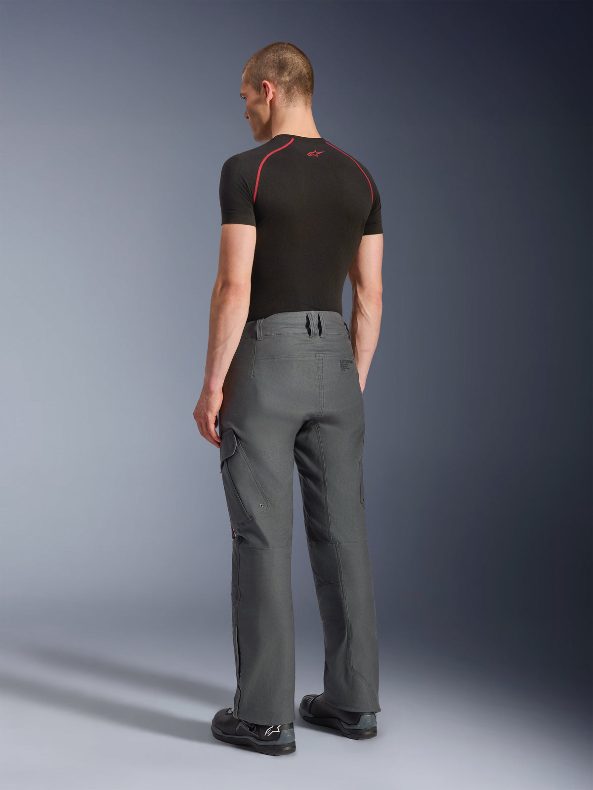 Alpinestars Borrego Drystar® Pants, calças de motociclismo de aventura, cinzento antracite, vista traseira, com aspeto de lona encerada e bolsos cargo, combinadas com uma t-shirt preta e botas de condução, design durável para estrada e touring de aventura
