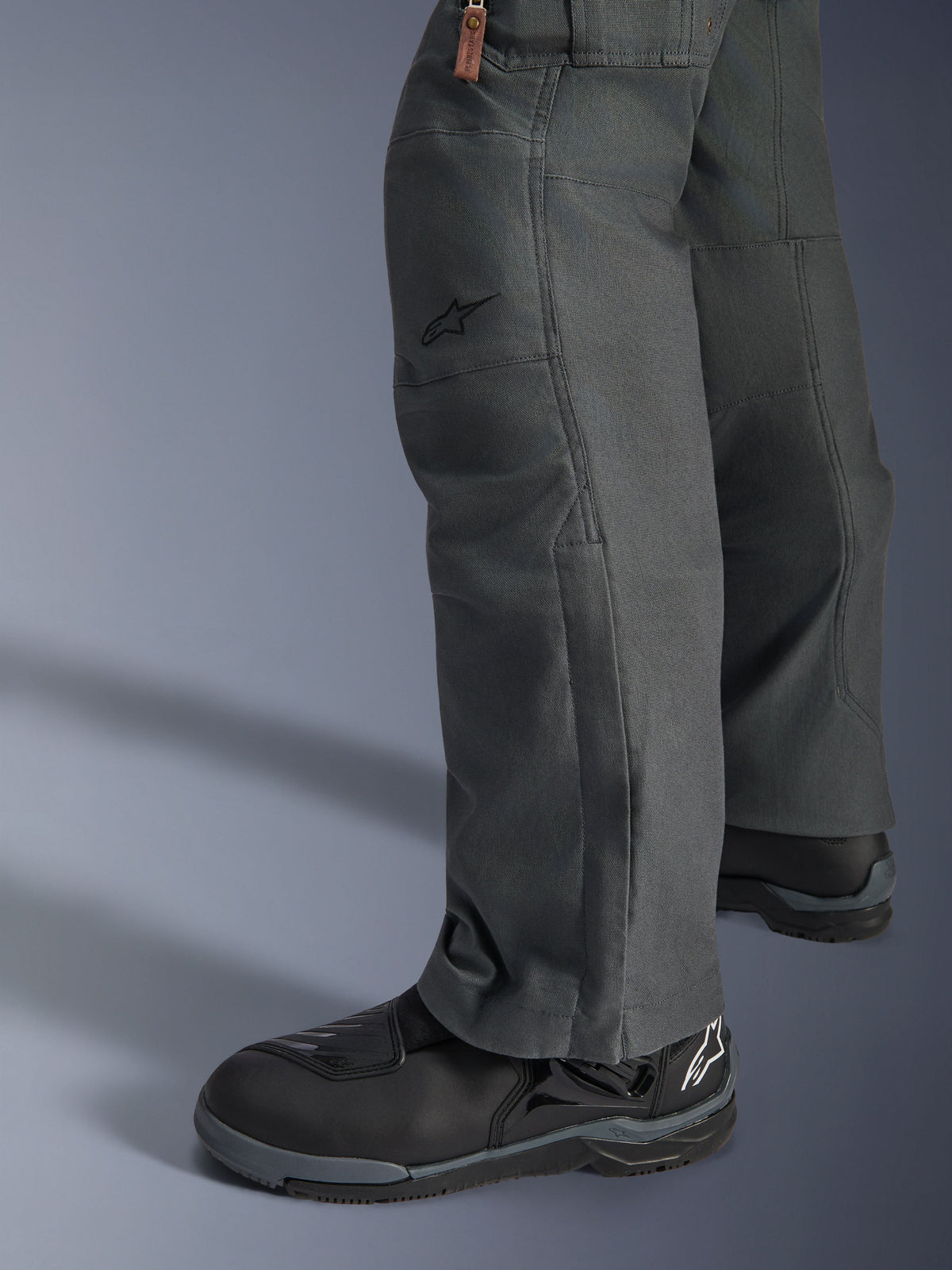Calças Alpinestars Borrego Drystar®, calças de motociclismo de aventura em cinzento antracite com aparência de lona encerada, construção têxtil vista de lado com botas de condução pretas, design robusto para estrada e touring de aventura