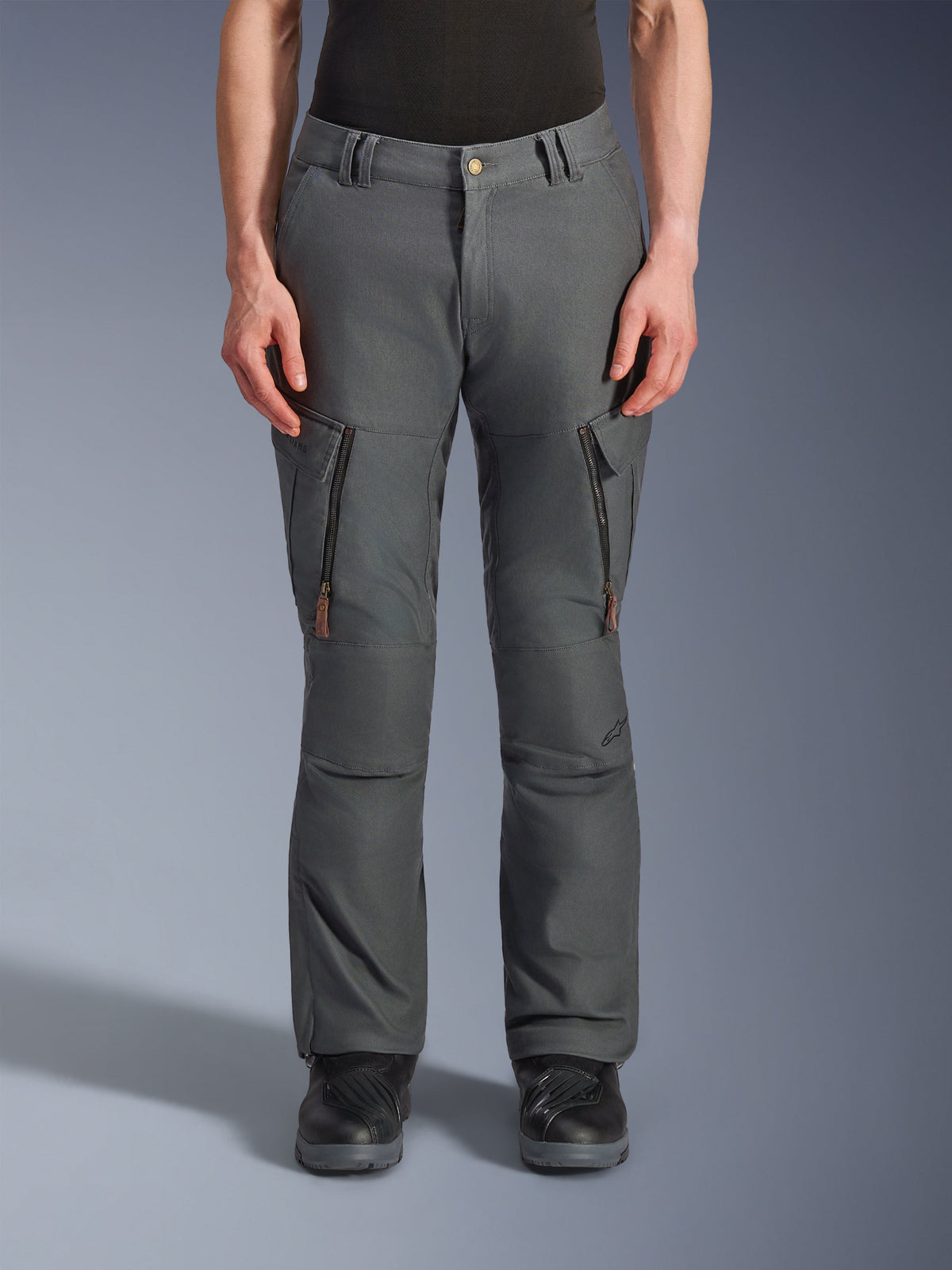 Alpinestars Borrego Drystar® Pants, calças de motociclismo de aventura, cinzento antracite, com bolsos verticais com fecho de correr nas coxas e puxadores em pele, fecho de botão na cintura e passadores de cinto, usadas por um modelo com botas de motociclismo pretas