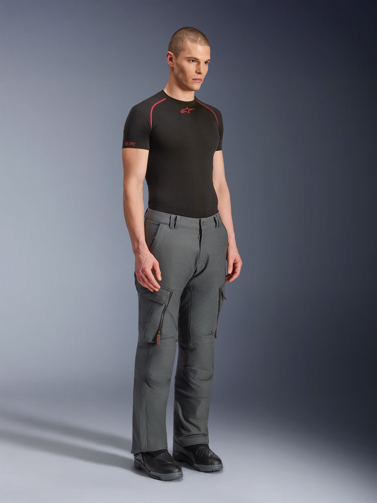 Modelo masculino a usar Calças Alpinestars Borrego Drystar®, calças de motociclismo de aventura em cinzento antracite, design inspirado em vestuário de trabalho com bolsos cargo e aberturas de ventilação com fecho, combinadas com uma camisola interior de manga curta preta e botas pretas