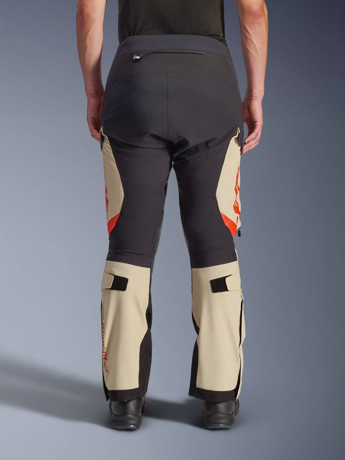 Vista traseira das calças Alpinestars Halo Pro Drystar® XF Laminated, em Mastic Black com painéis bege e detalhes em laranja, calças de motociclismo de aventura com construção têxtil técnica, usadas com botas de condução pretas.