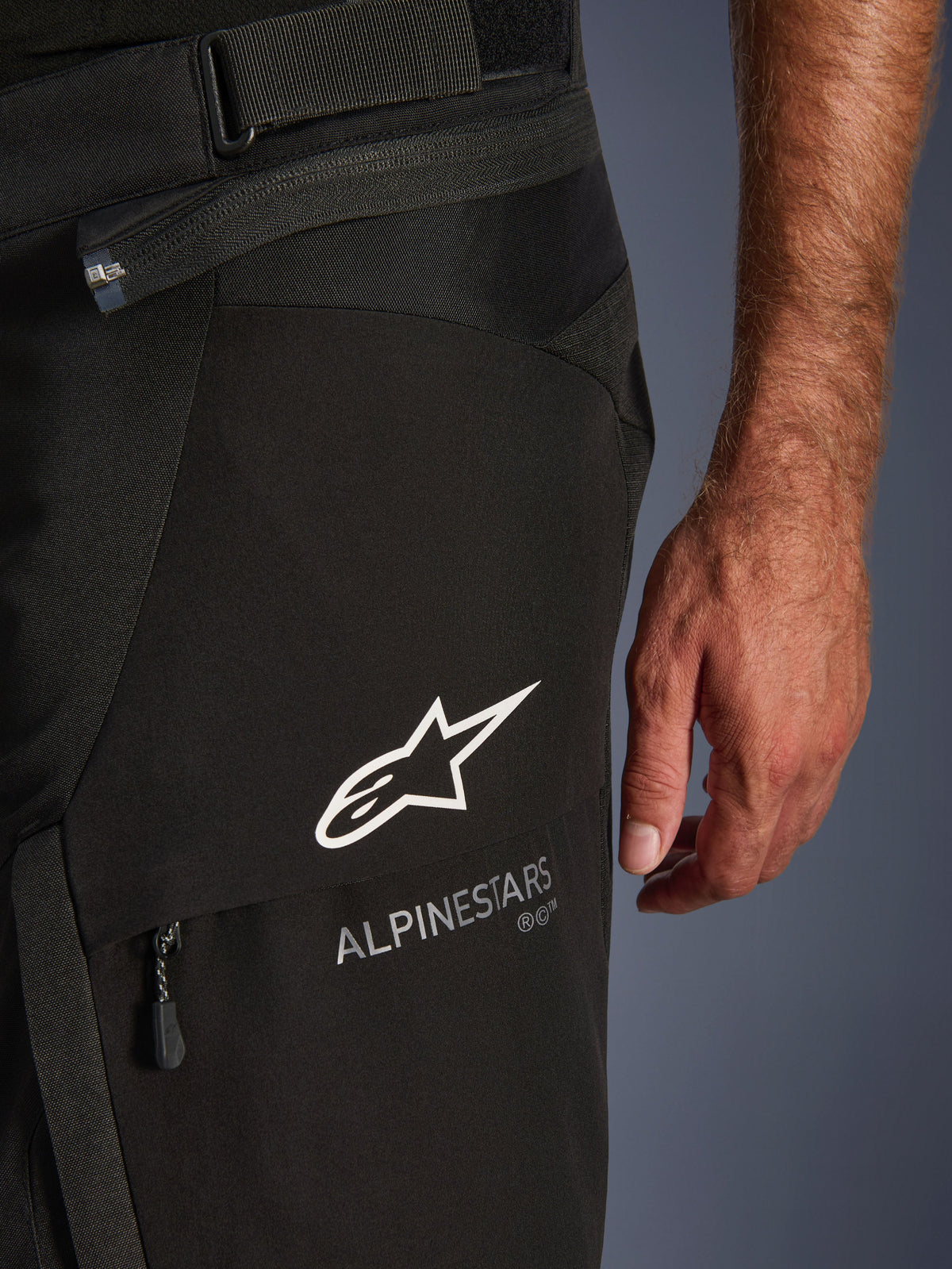 Alpinestars Andes V4 Drystar® Pants, Close-up detail of black motorcycle pants, featuring white Alpinestars logo and text branding on the thigh, showing technical fabric textures, zipper, and adjustable waist strap, adventure touring gear