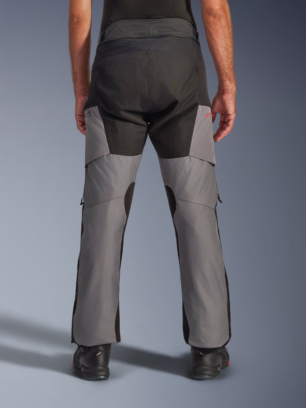 Alpinestars Andes V4 Drystar® Pants, calças de motociclismo Adventure, pretas e cinzento escuro, vista traseira apresentando o assento reforçado a preto e pernas cinzentas, com um pequeno logótipo vermelho, combinadas com botas de condução pretas, construção têxtil durável para touring em todas as condições climatéricas.