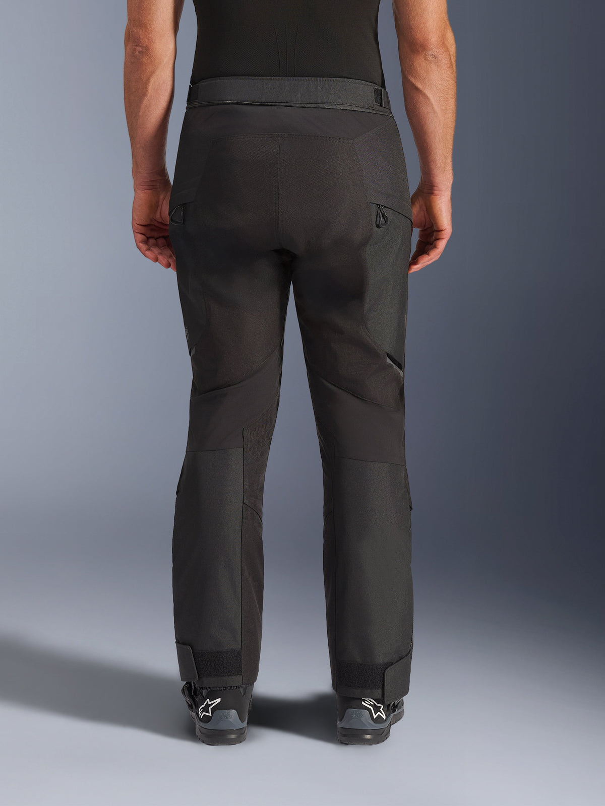Calças Alpinestars Cusco Drystar®, calças de motociclismo, pretas, vista traseira vestidas por um motociclista, com painéis têxteis texturizados, aberturas de ventilação com fecho nas coxas e fechos ajustáveis nos tornozelos, combinadas com botas Alpinestars pretas.