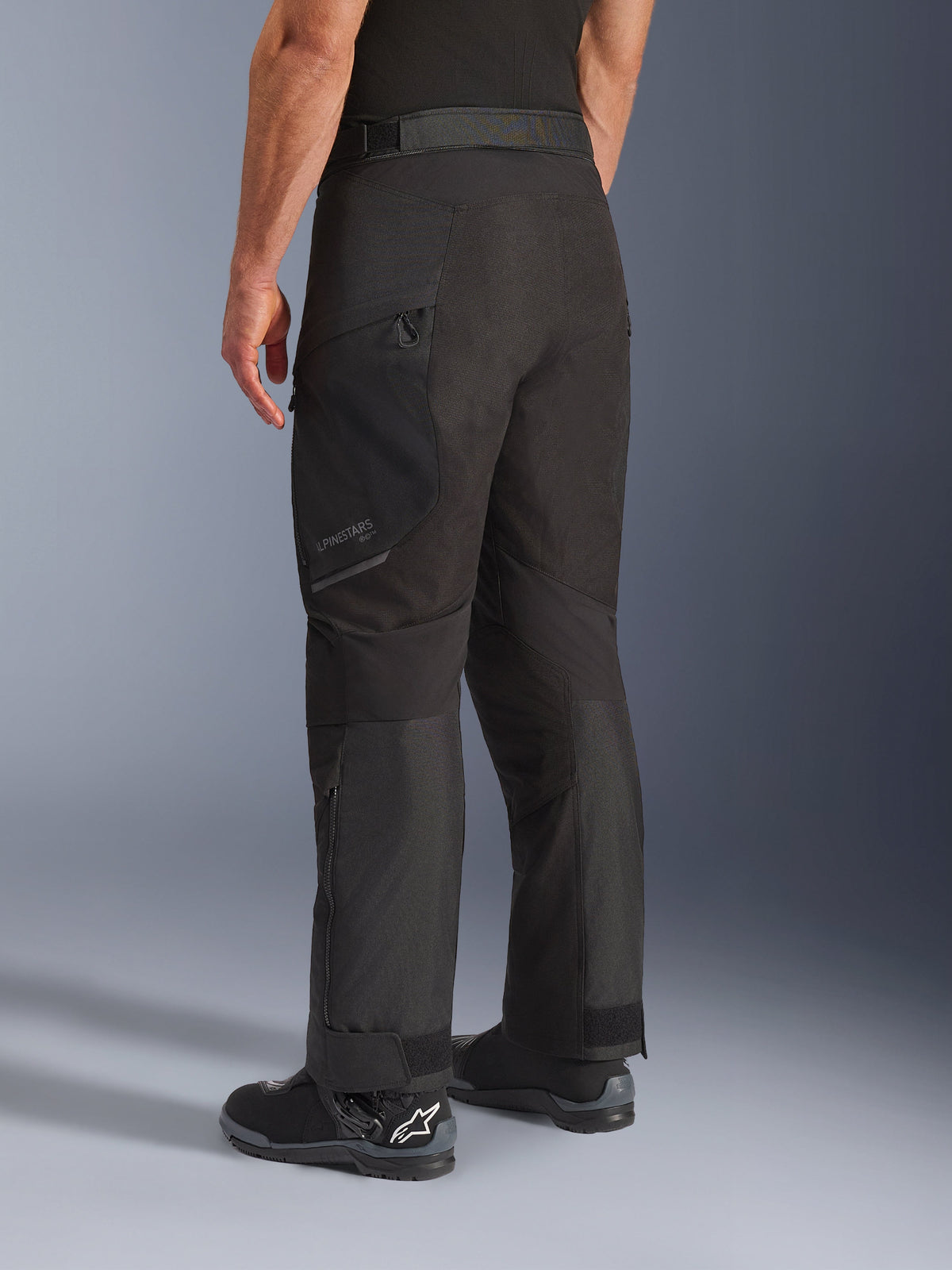 Calças Alpinestars Cusco Drystar®, calças de motociclismo, pretas, vista traseira em três quartos mostrando o bolso na coxa com fecho de correr e painéis articulados, usadas com botas de motociclismo pretas, design durável de turismo de aventura com branding subtil