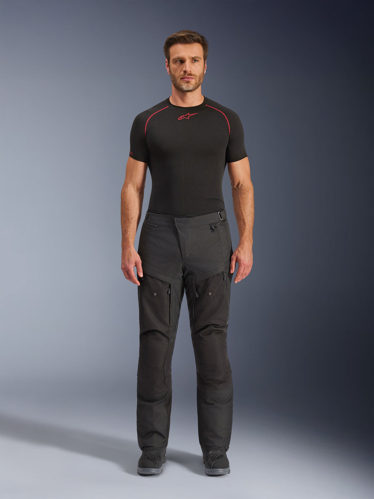 Calças Alpinestars Cusco Drystar®, calças de motociclismo em têxtil, pretas, vestidas por um modelo com uma camada de base preta e botas, design de turismo de aventura com painéis de ventilação com fecho éclair nas coxas e construção durável multi-painel