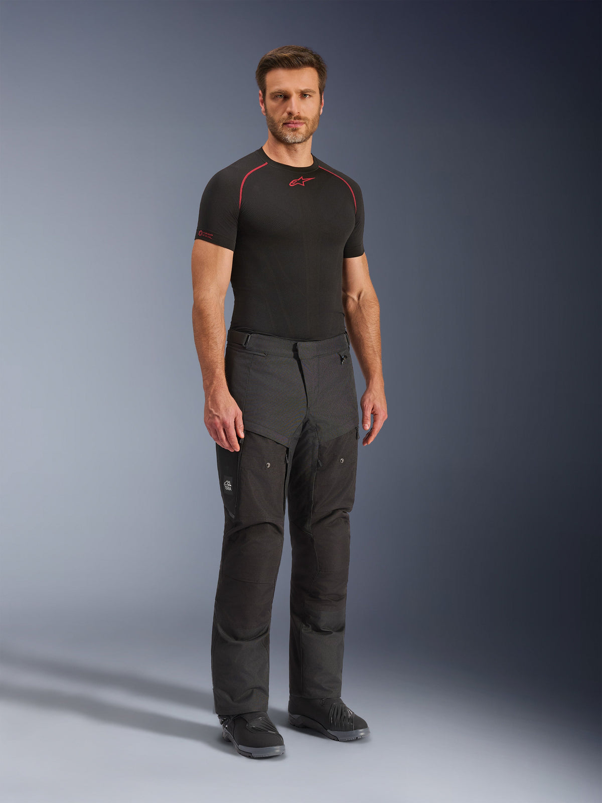 Calças Alpinestars Cusco Drystar®, calças de motociclismo em tecido pretas, vestidas por um modelo com uma base layer preta e botas, com tecido ripstop, bolsos cargo grandes com a marca 'Terra' e design ergonómico para condução de aventura.