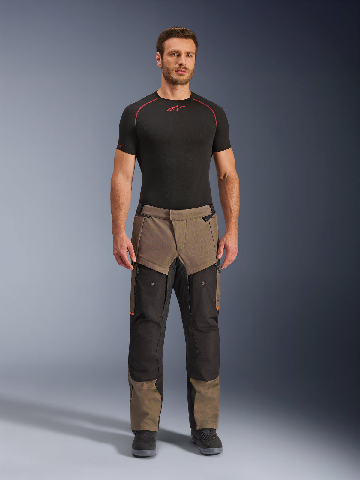 Alpinestars Cusco Drystar® Pants, Walnut Black, calças de motociclismo para adventure touring, vestidas por um modelo masculino com uma camada de base preta e botas pretas, com painéis em walnut e preto, detalhes em laranja e fechos de ventilação direta nas coxas.