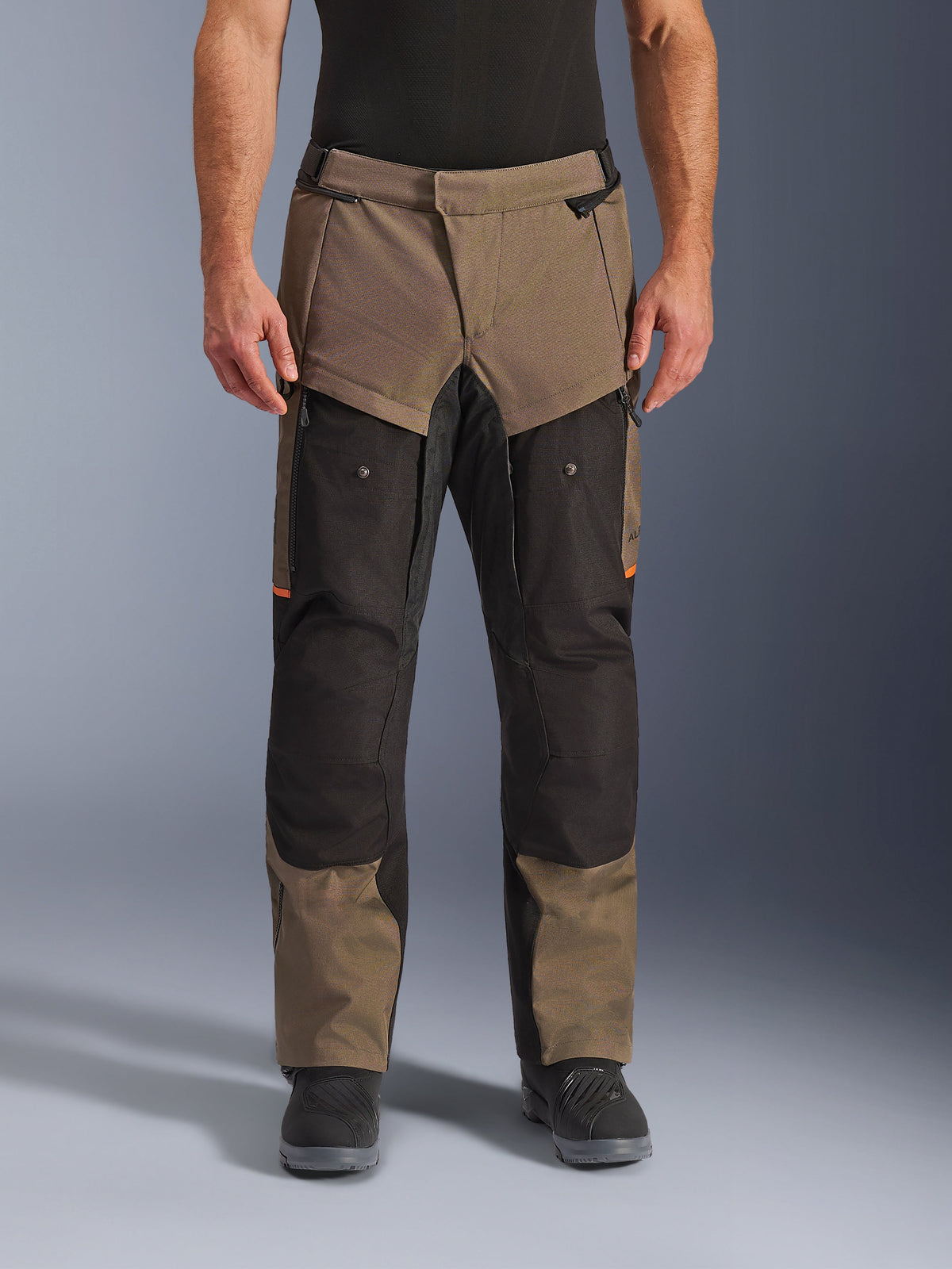 Calças Alpinestars Cusco Drystar®, calças de motociclismo adventure touring, walnut e preto com pormenores cor de laranja, vista frontal num modelo, construção em tecido técnico, bolsos cargo e fechos de ventilação, usadas com botas de motociclismo pretas