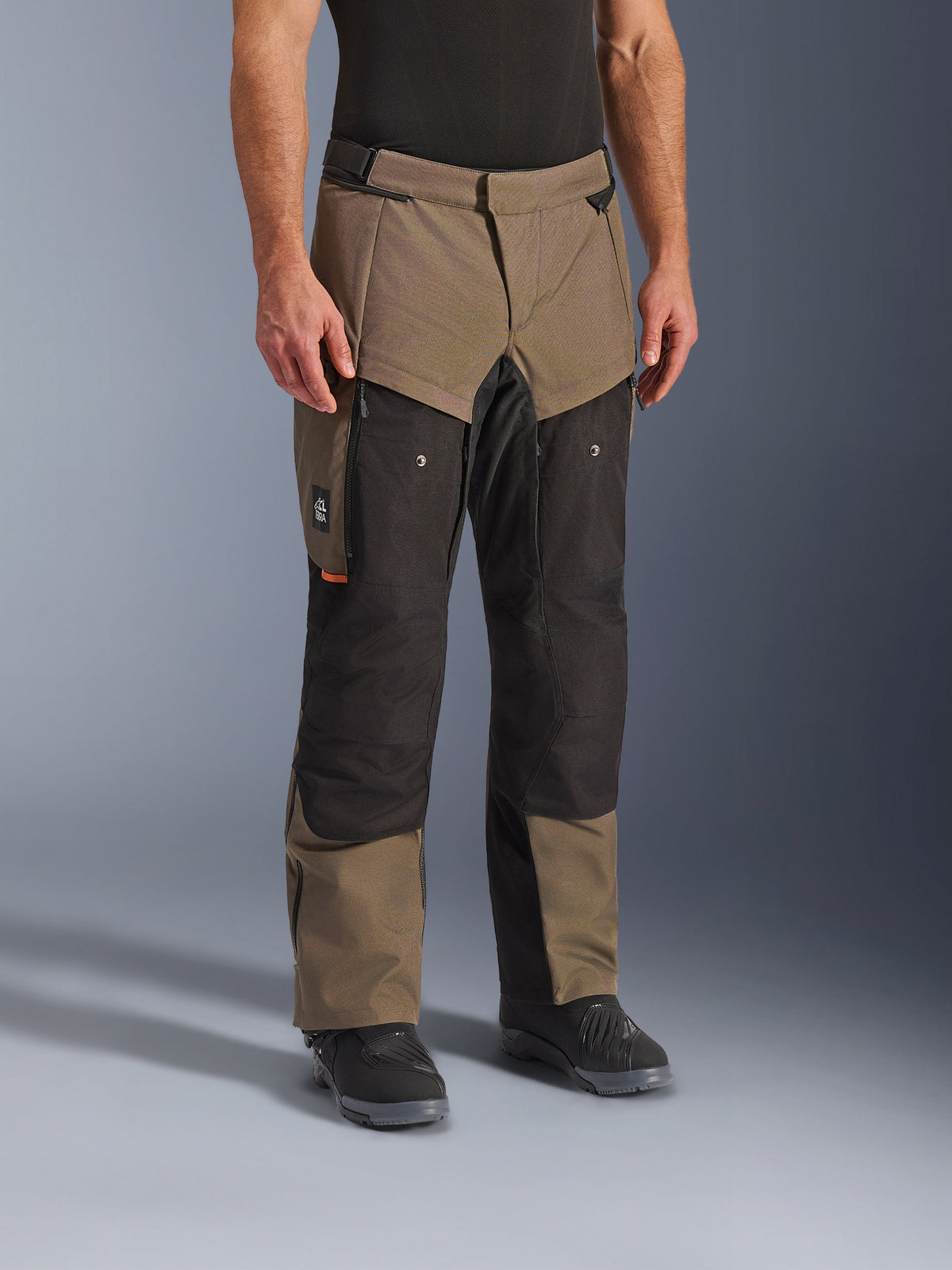 Calças Alpinestars Cusco Drystar®, calças de motociclismo de aventura, Walnut Black, apresentadas num modelo com botas pretas, com um design multi-painel em castanho e preto, bolsos com fecho e joelhos reforçados para turismo
