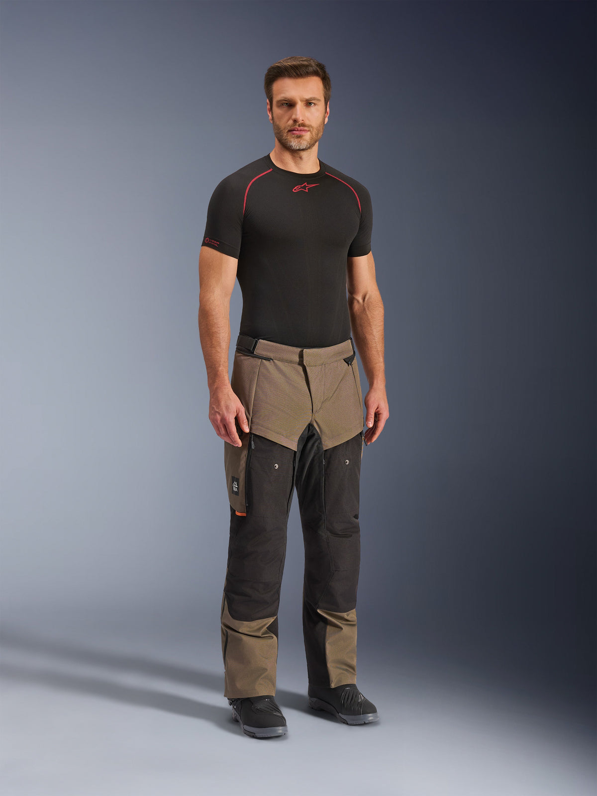 Calças Alpinestars Cusco Drystar®, calças de motociclismo de aventura, em nogueira e preto, usadas por um modelo com uma base layer preta e botas, com bolsos cargo grandes nas coxas, fechos de ventilação e um design robusto em dois tons