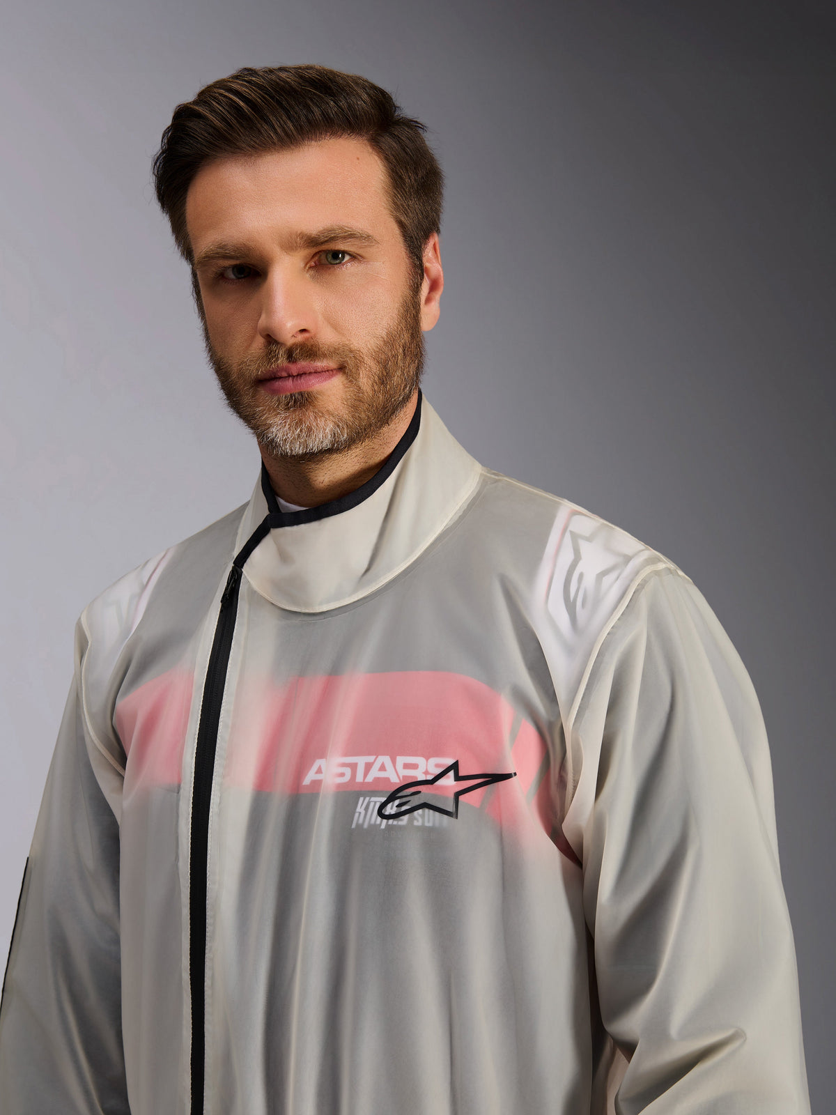 Macacão de Chuva para Kart Alpinestars, sobrefato de chuva para karting, transparente, usado por um homem, material impermeável transparente que deixa ver detalhes do fato de corrida vermelho e branco por baixo, gola alta com fecho preto e logótipo da estrela Alpinestars no peito